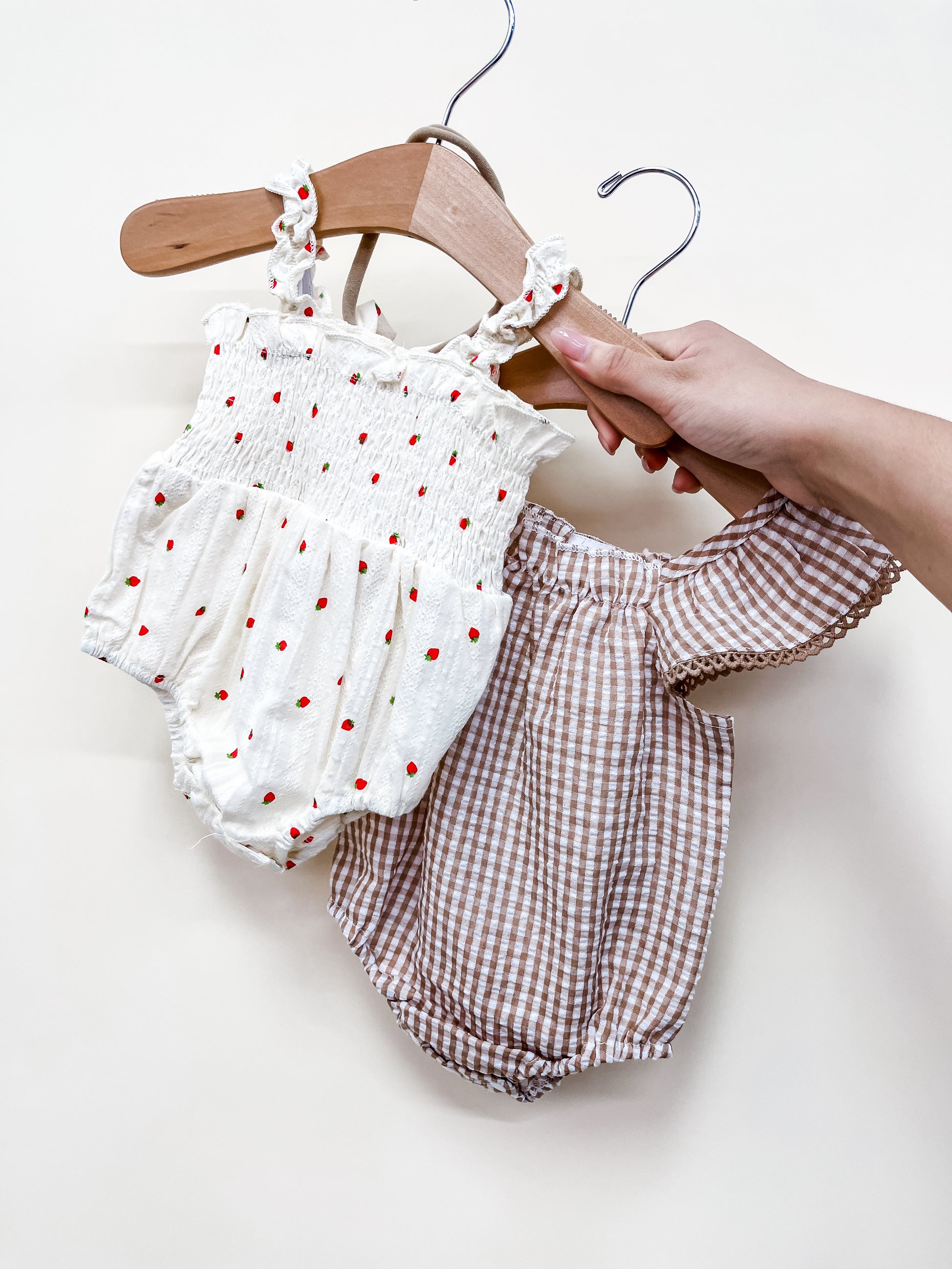 A hand displays the Tiny Tot Bundles "Bundle Of The Day - Girls": two baby rompers—one white with red polka dots and smocking, the other brown and white gingham with puffed sleeves—on wooden hangers against a plain white background.