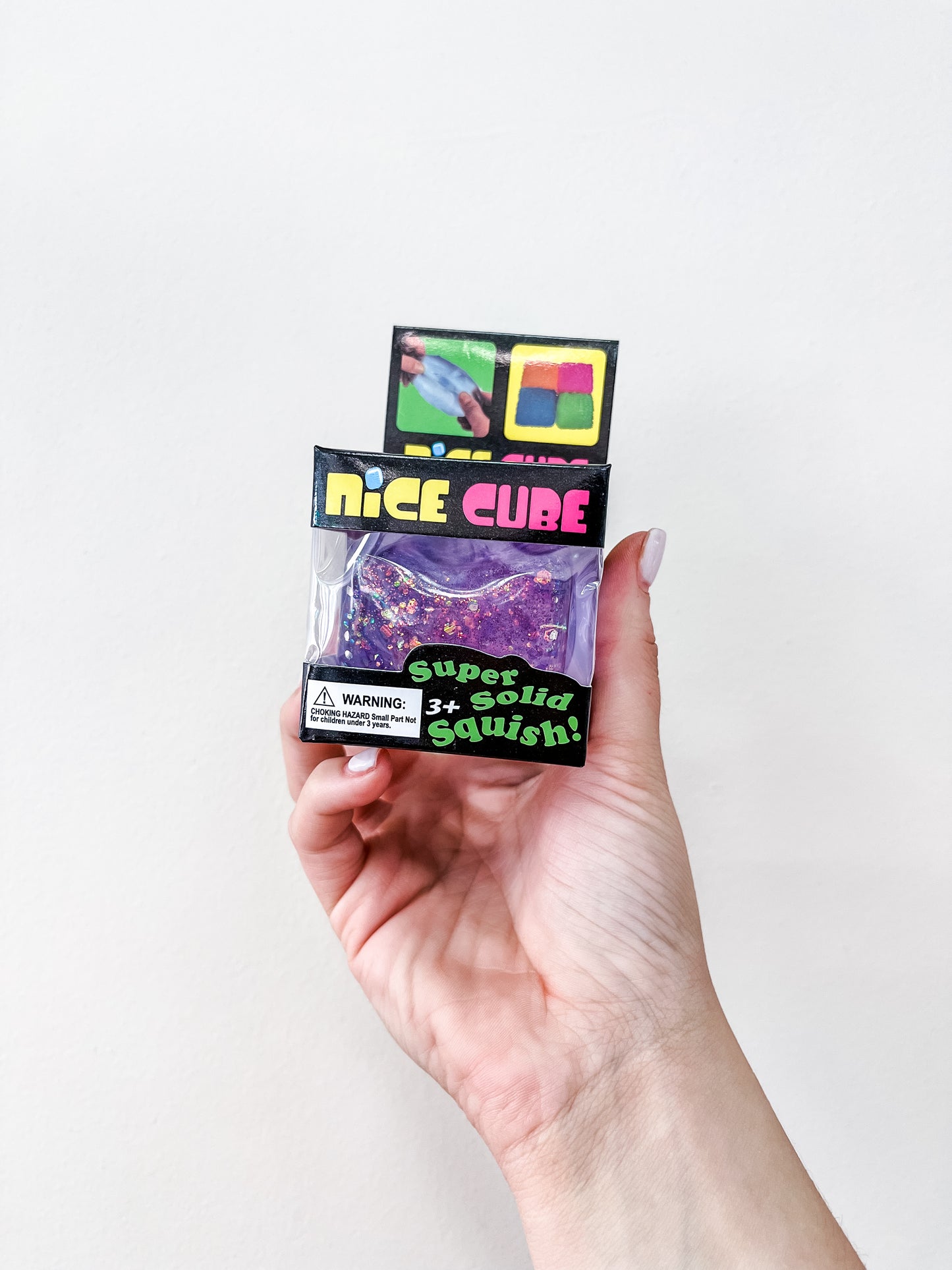 A hand holds a small Prime Opticals Glitter Squishy Cube box with a purple, sparkly toy inside. The packaging says "Super Solid Squish!", shows a child playing, and has a warning label at the bottom left.