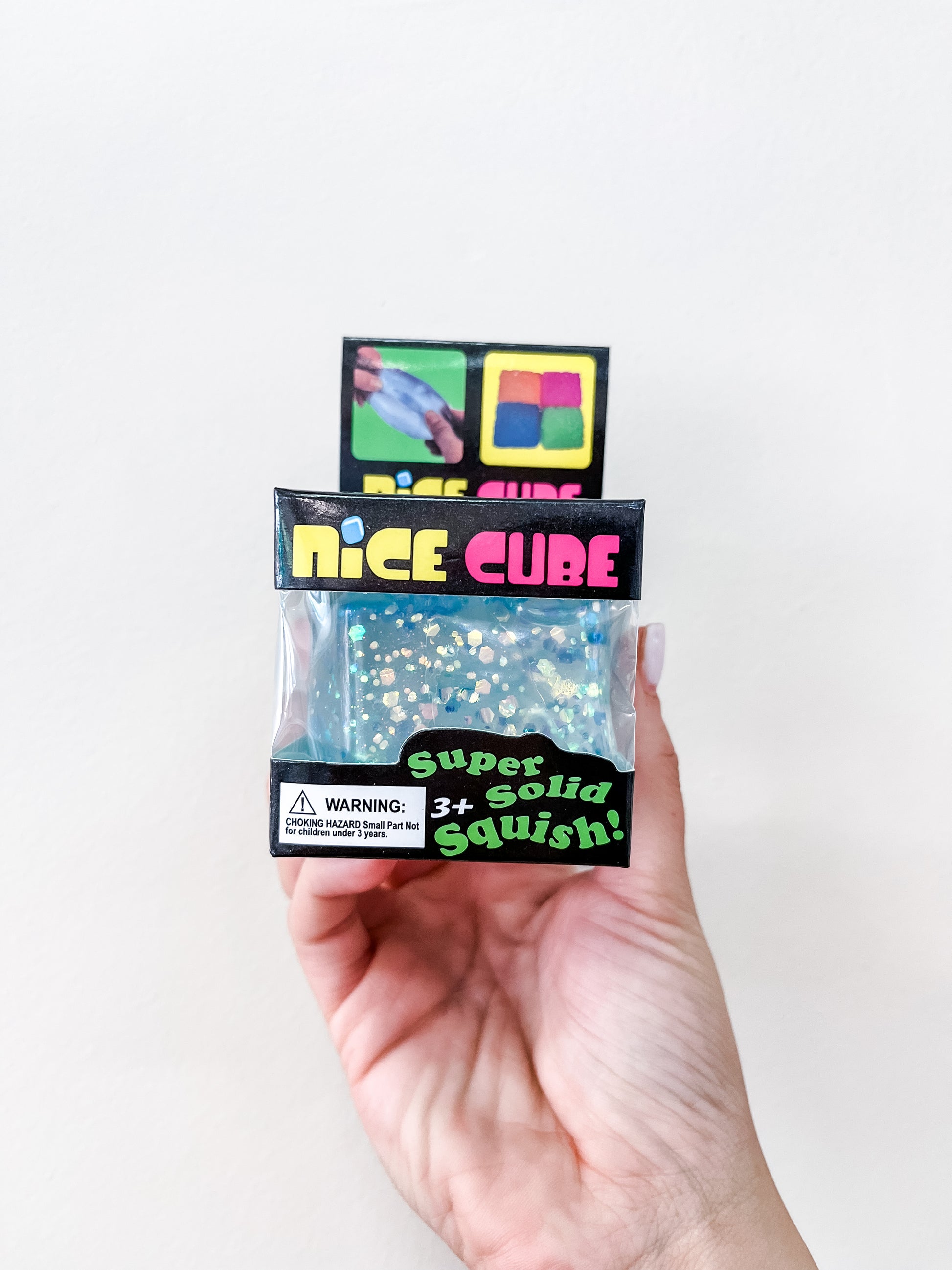 A hand holds a packaged Prime Opticals Glitter Squishy Cube—a sparkly blue toy with a "for ages 3 and up" warning—against a plain light wall.