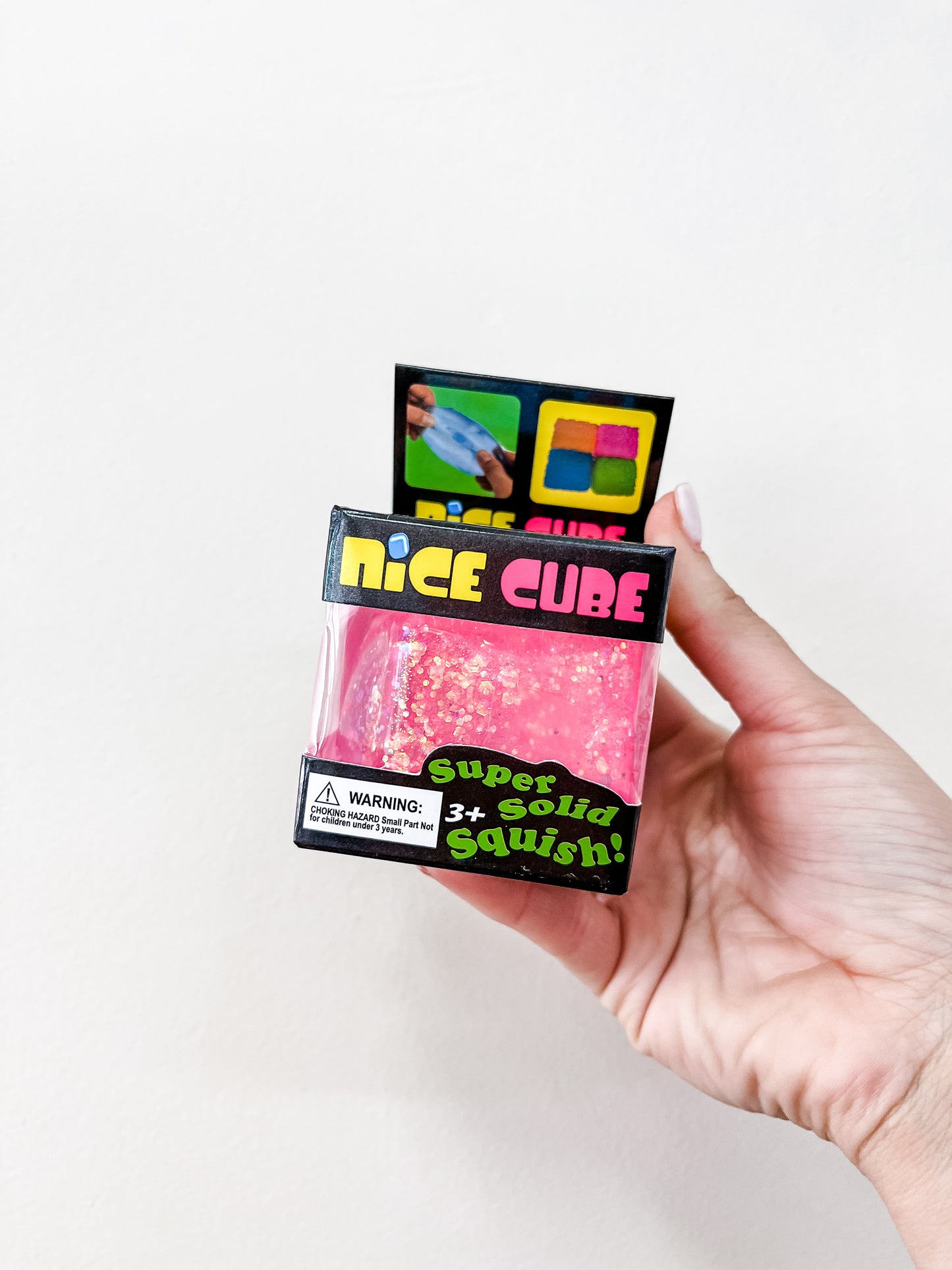 A hand holds a pink Glitter Squishy Cube by Prime Opticals, still in its packaging labeled "Super Solid Squish! 3+", set against a plain white background.