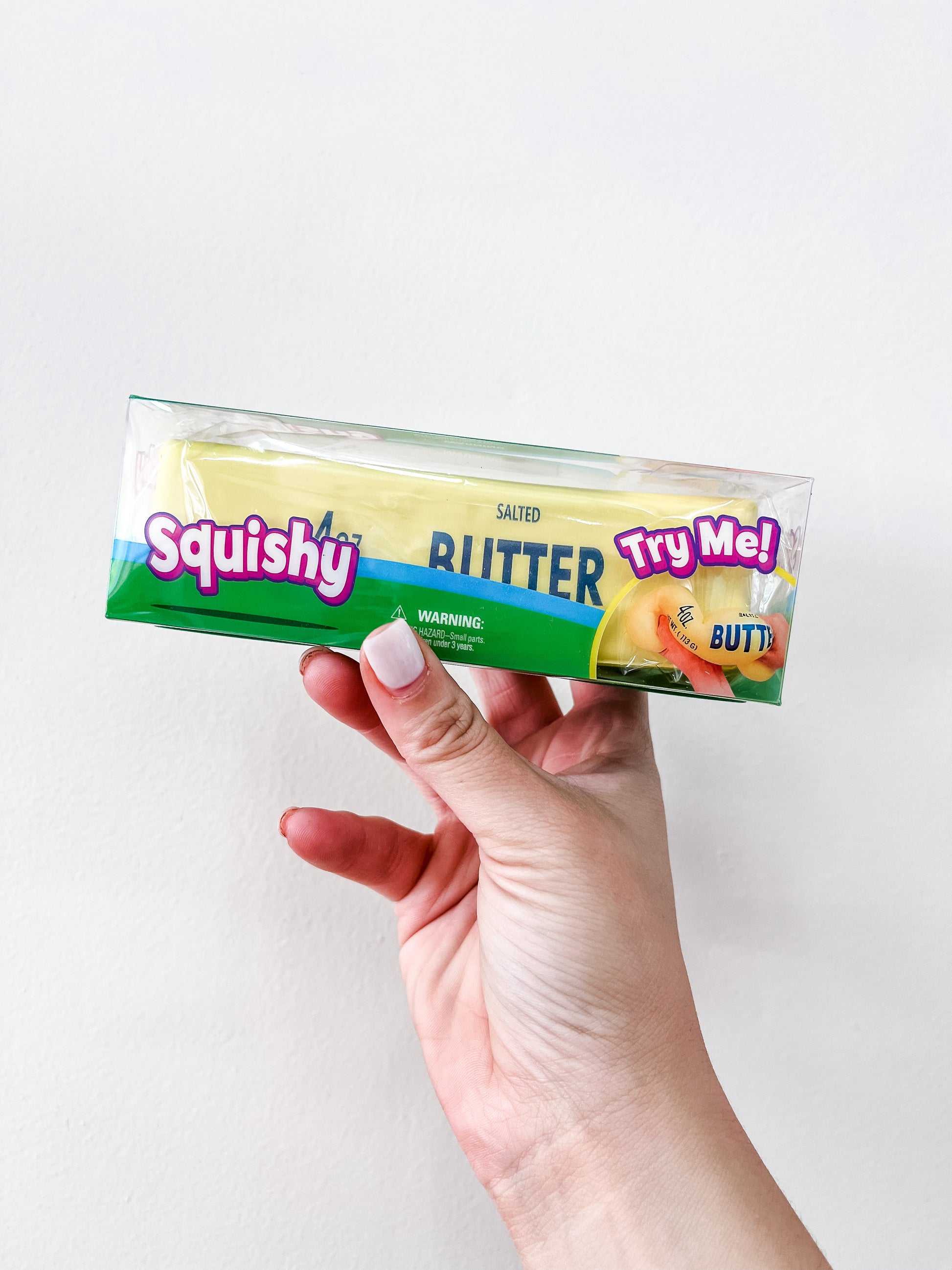 A hand holds a Boxed Yellow Butter Squishy - 4oz by Prime Opticals, packaged to resemble a stick of salted butter with colorful "Squishy" and "Try Me!" text, set against a plain white background.