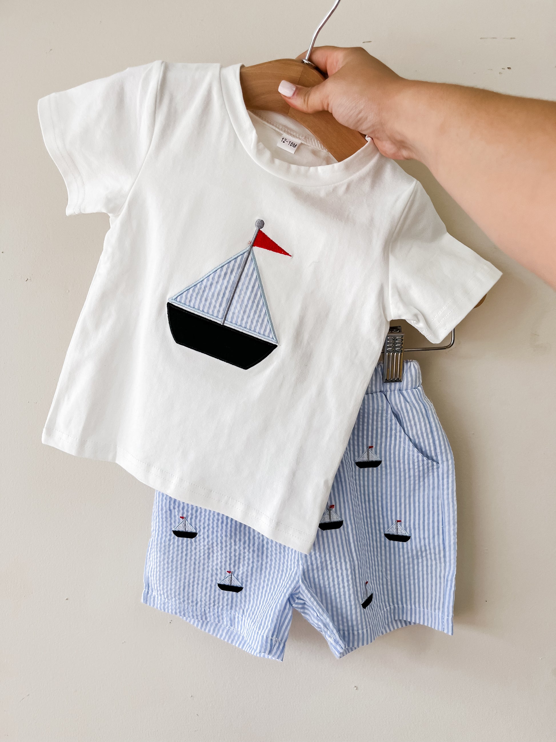 A hand holds the Tiny Tot Bundles Stripe Sailboat Short Set, featuring a white t-shirt with a sailboat graphic and matching blue-and-white striped shorts adorned with small sailboat prints.