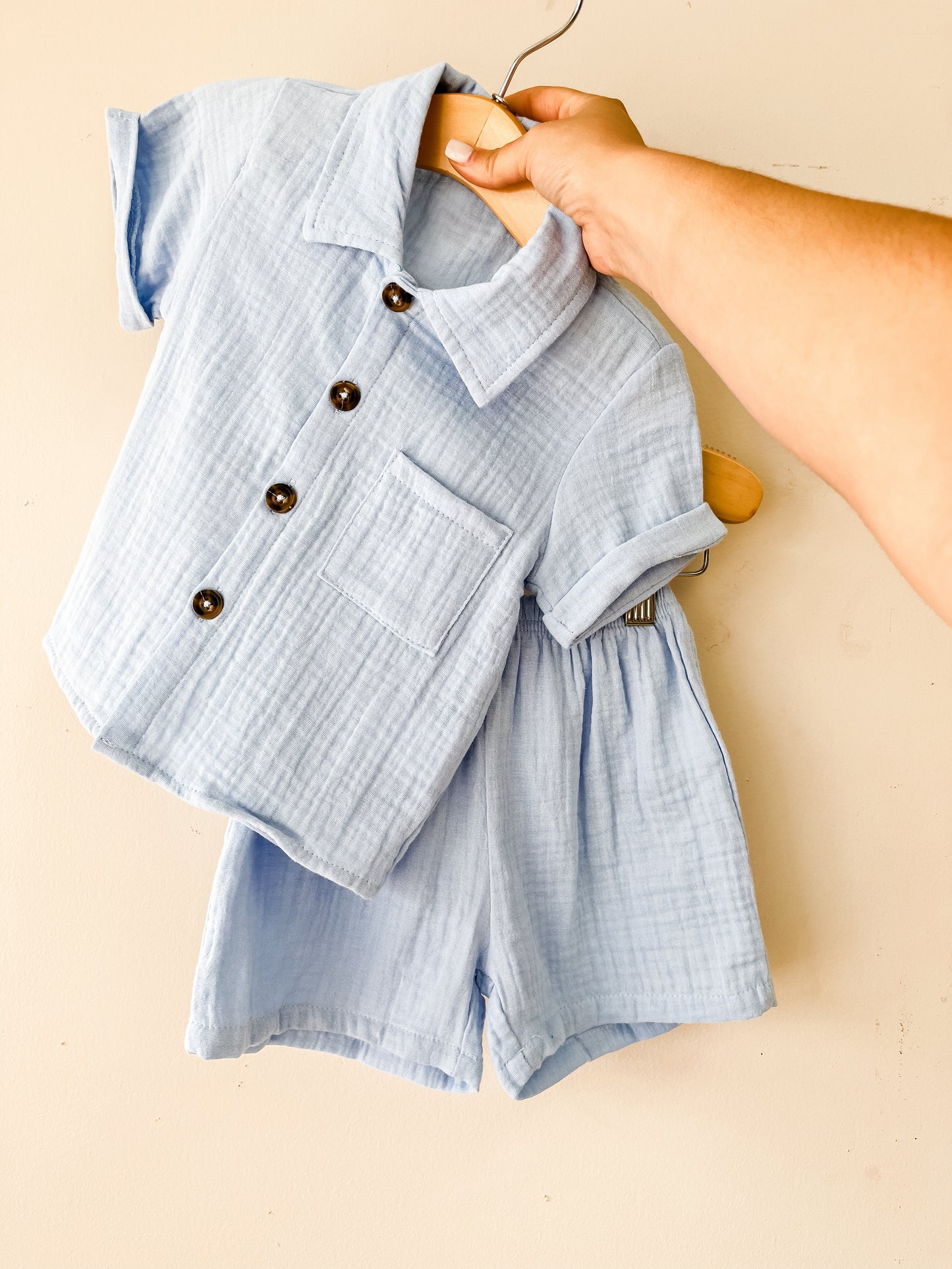 A hand holds up the Blue Linen Set by Tiny Tot Bundles, featuring a light blue, short-sleeved button-up shirt and matching shorts for children, both crafted from soft textured fabric against a plain light background.