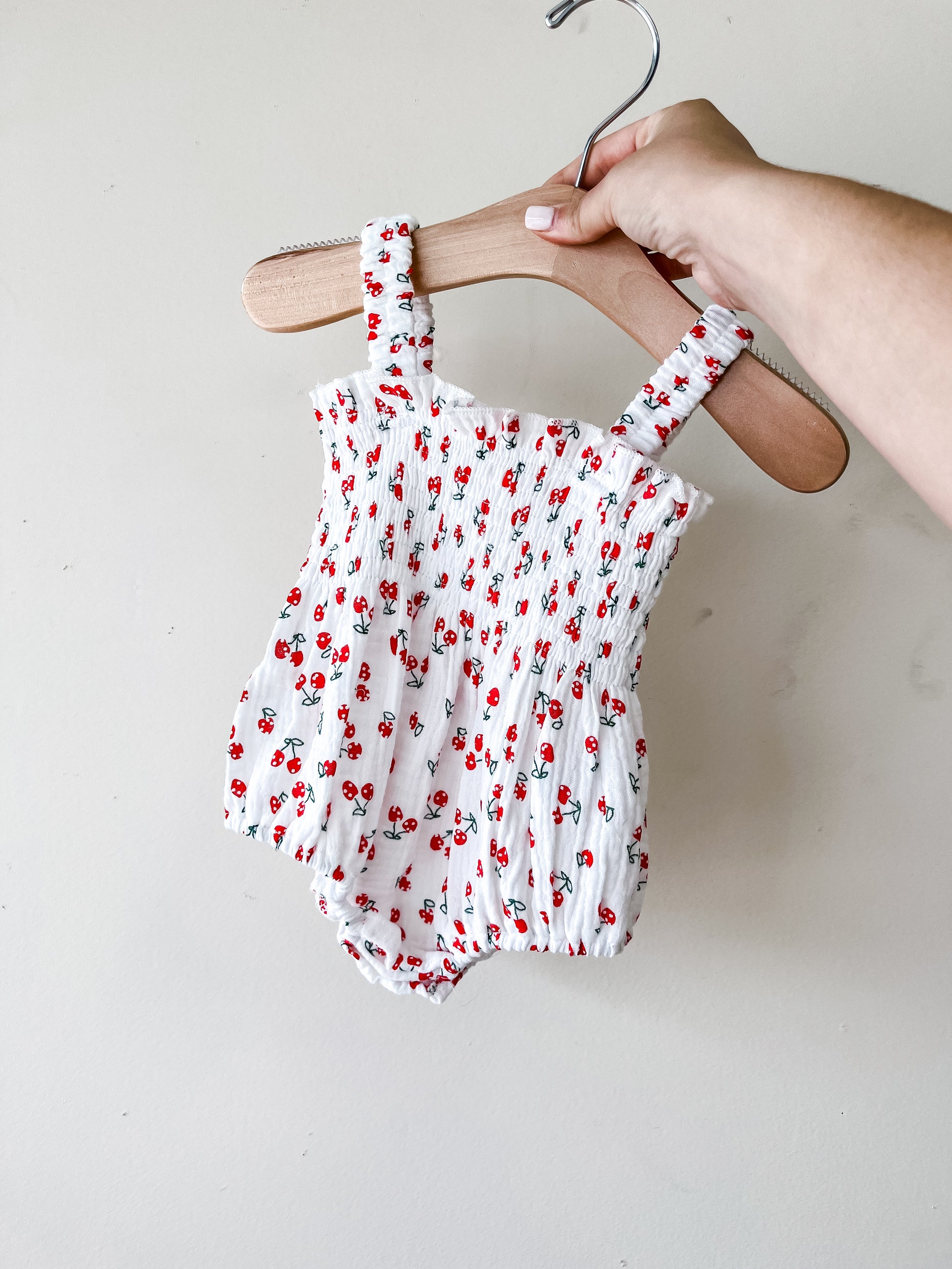 A hand holds a wooden hanger with the Tiny Tot Bundles Cherry Linen Romper, a white baby outfit adorned with red and green floral prints, set against a plain light background.