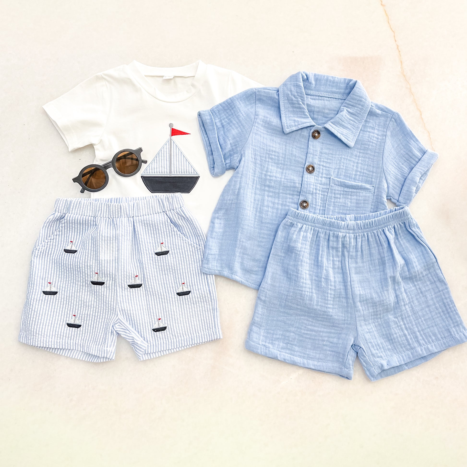 Flat lay of Bundle Of The Day - Boys by tiny: a white sailboat graphic tee, blue striped boat shorts, light blue button-up shirt with matching shorts, and round sunglasses.