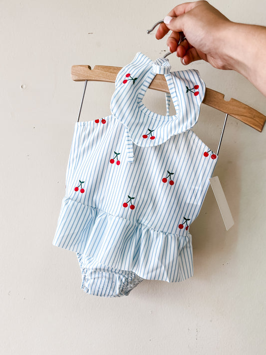A hand holds a hanger with the Tiny Tot Bundles Cherry Stripe Swimsuit, a baby girls' blue and white striped romper featuring red cherry prints, ruffled collar, and ruffled bottom, displayed against a plain light-colored wall.