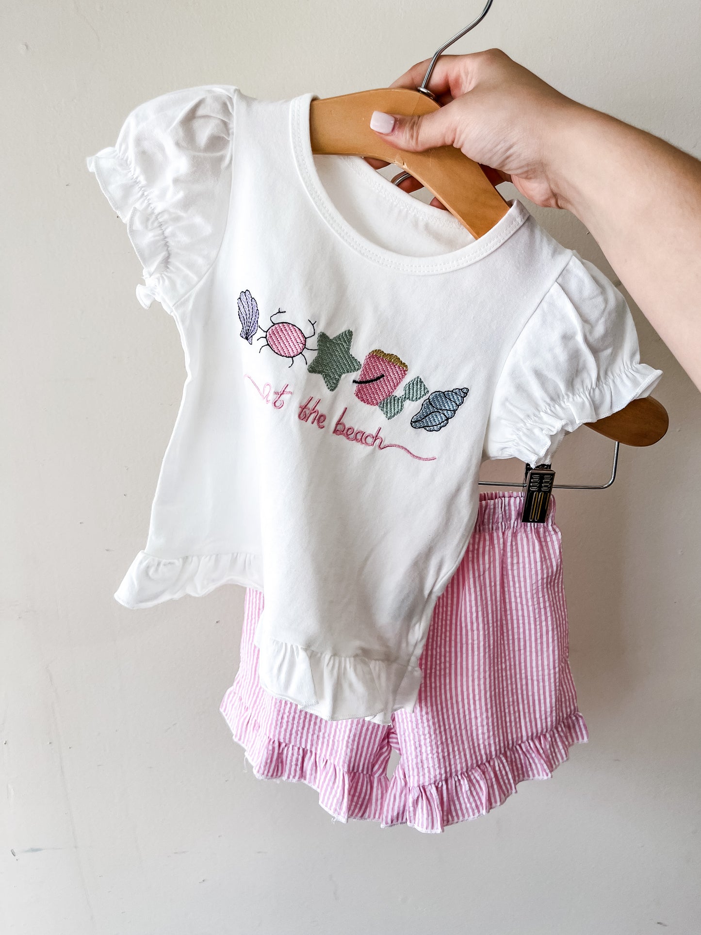 A hand holds the Tiny Tot Bundles At The Beach Pink Short Set: a white short-sleeve top with embroidered seashells and "at the beach" text, paired with pink-and-white striped ruffle shorts.