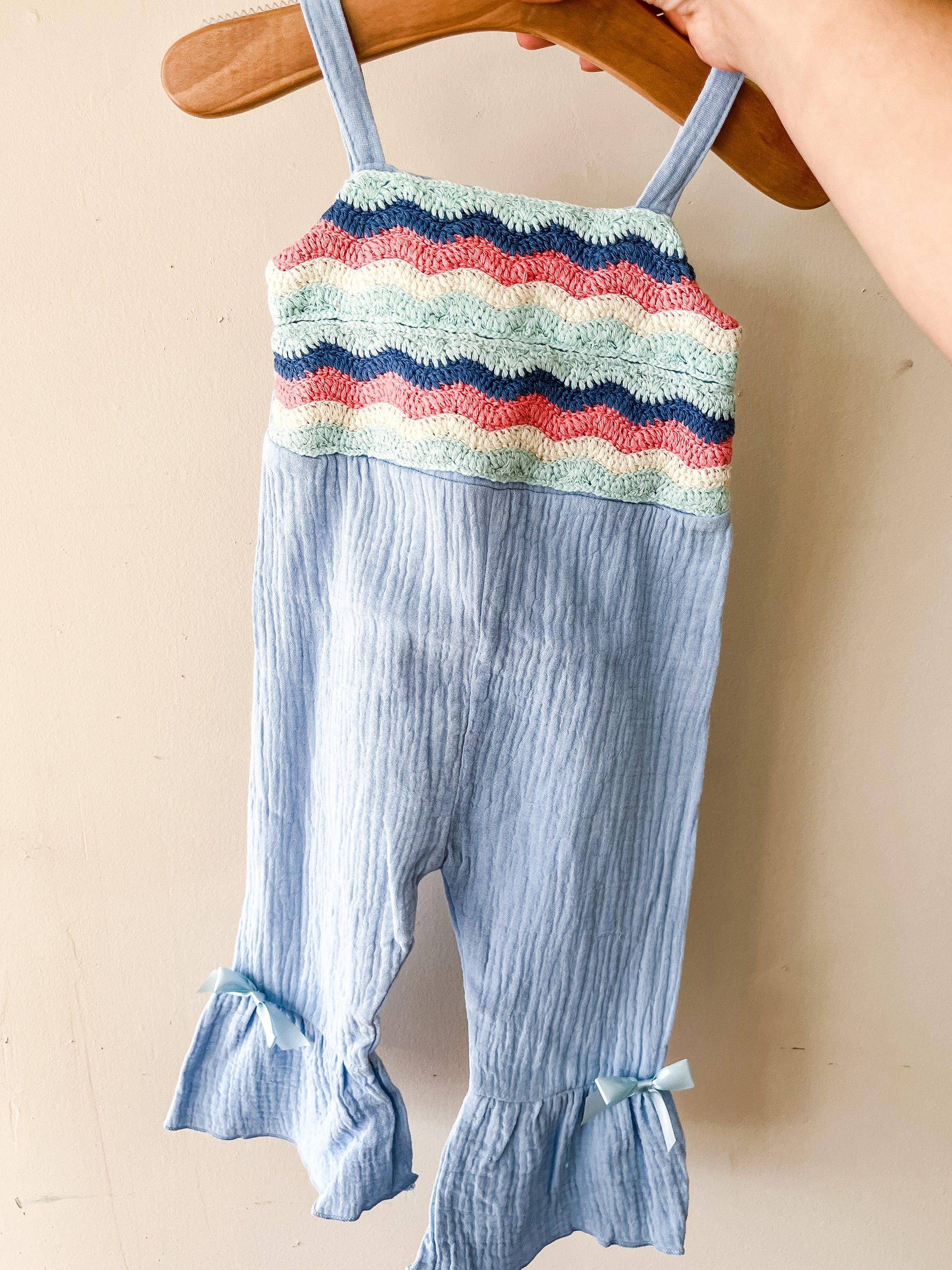 A hand holds the Seabreeze Ruffle Romper by Tiny Tot Bundles—a light blue toddler jumpsuit with ruffled legs, bow accents, and a textured top featuring mint green, pink, and navy zigzag stripes.