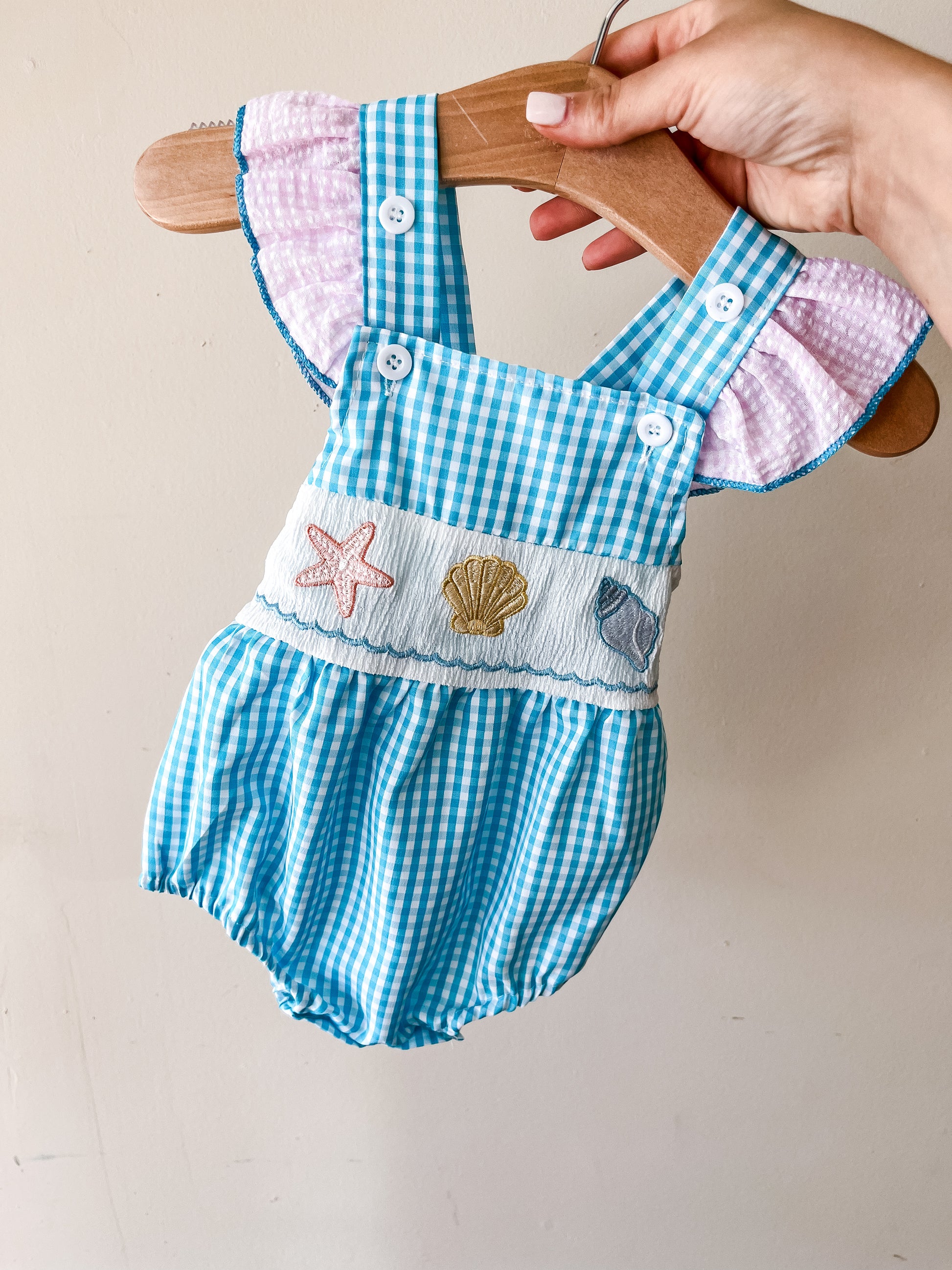 A hand holds Tiny Tot Bundles' Coastal Cutie Romper—a blue and white gingham baby outfit with pink ruffled sleeves and embroidered sea creatures (starfish, shell, fish) on a white band.