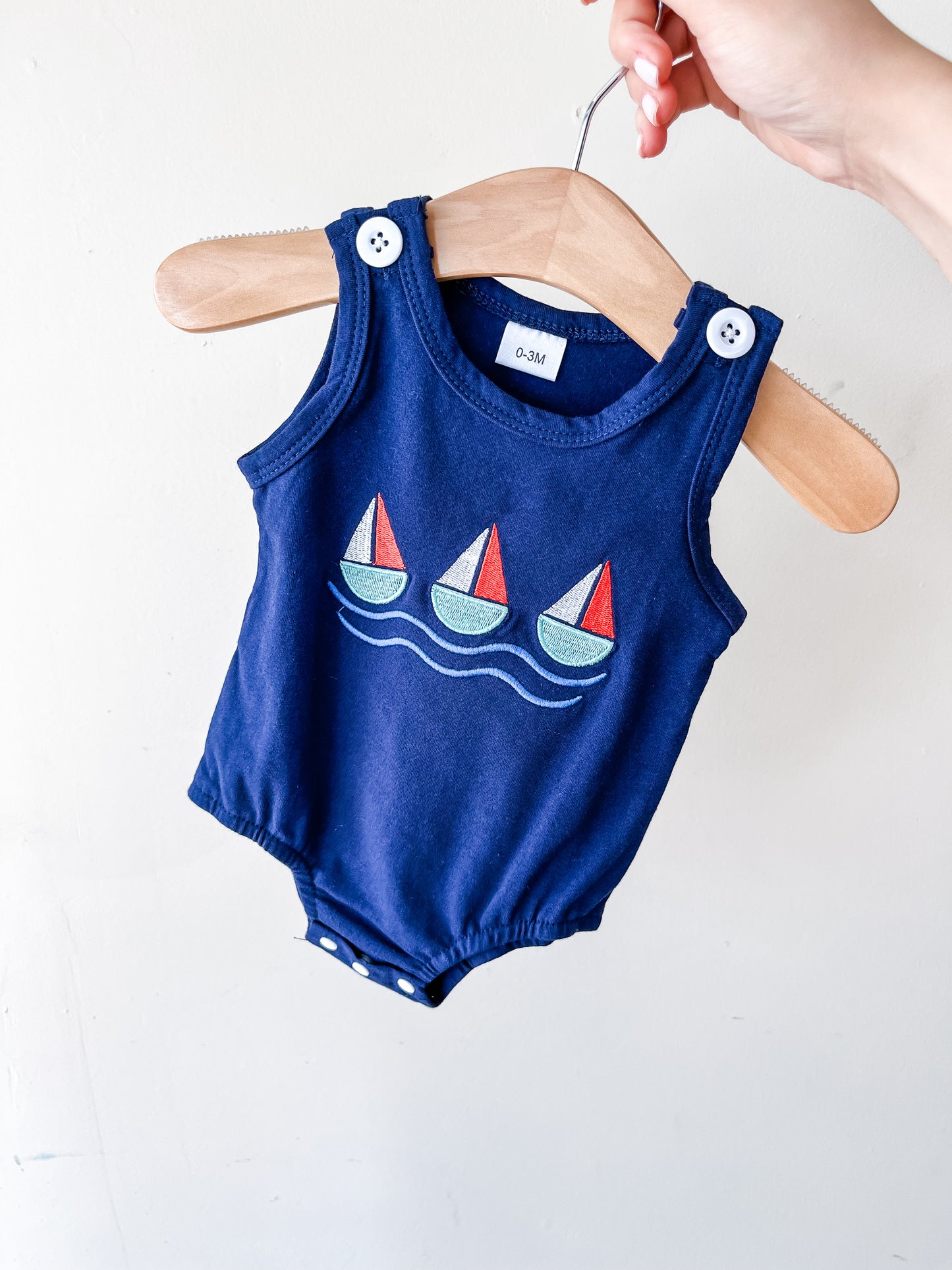 A hand holds a hanger displaying the Little Voyager Tank Romper by Tiny Tot Bundles—a navy blue sleeveless baby romper with white buttons and a sailboat and wave design on the front.