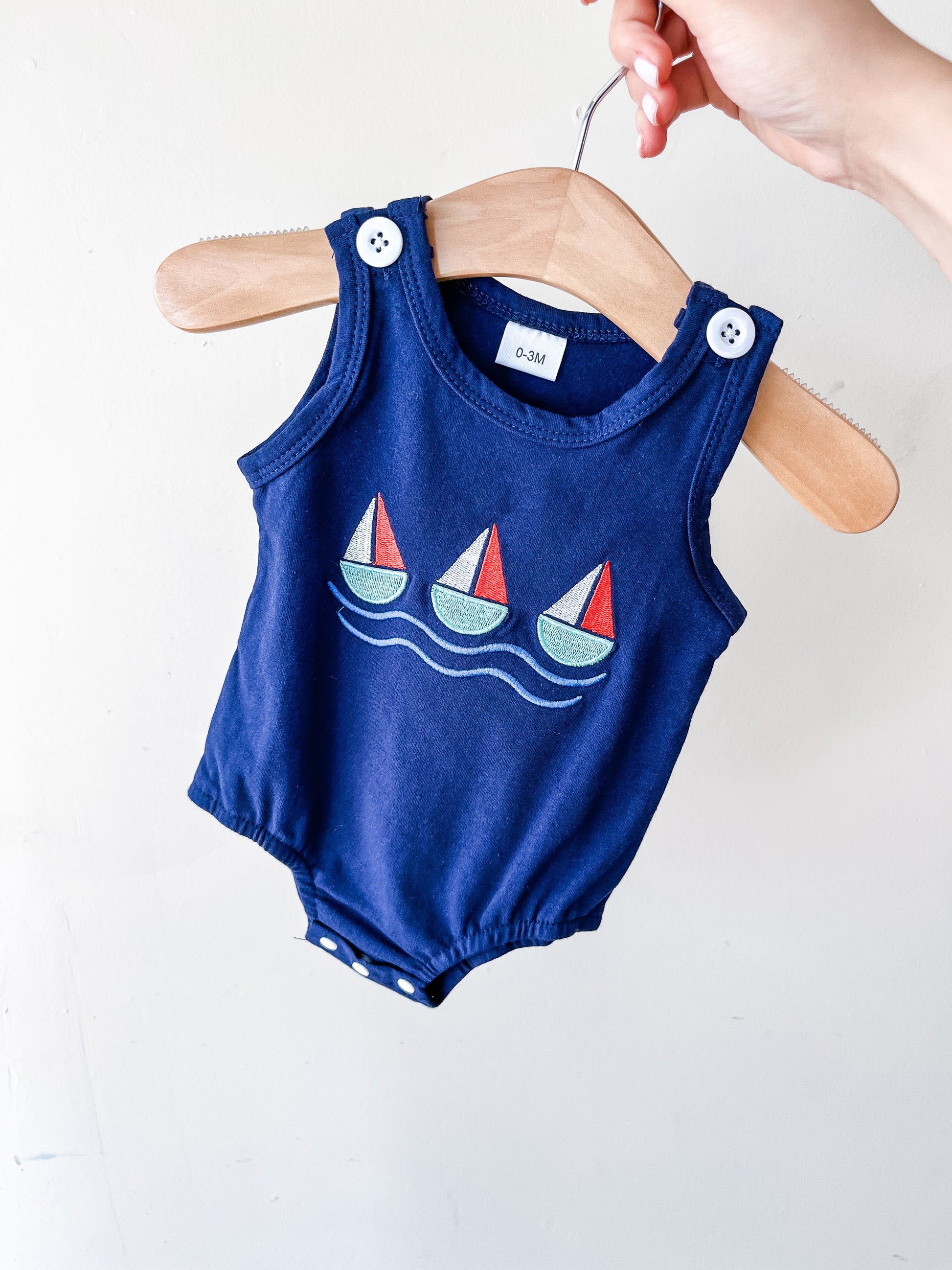 A hand holds a hanger displaying the Little Voyager Tank Romper by Tiny Tot Bundles—a navy blue sleeveless baby romper with white buttons and a sailboat and wave design on the front.
