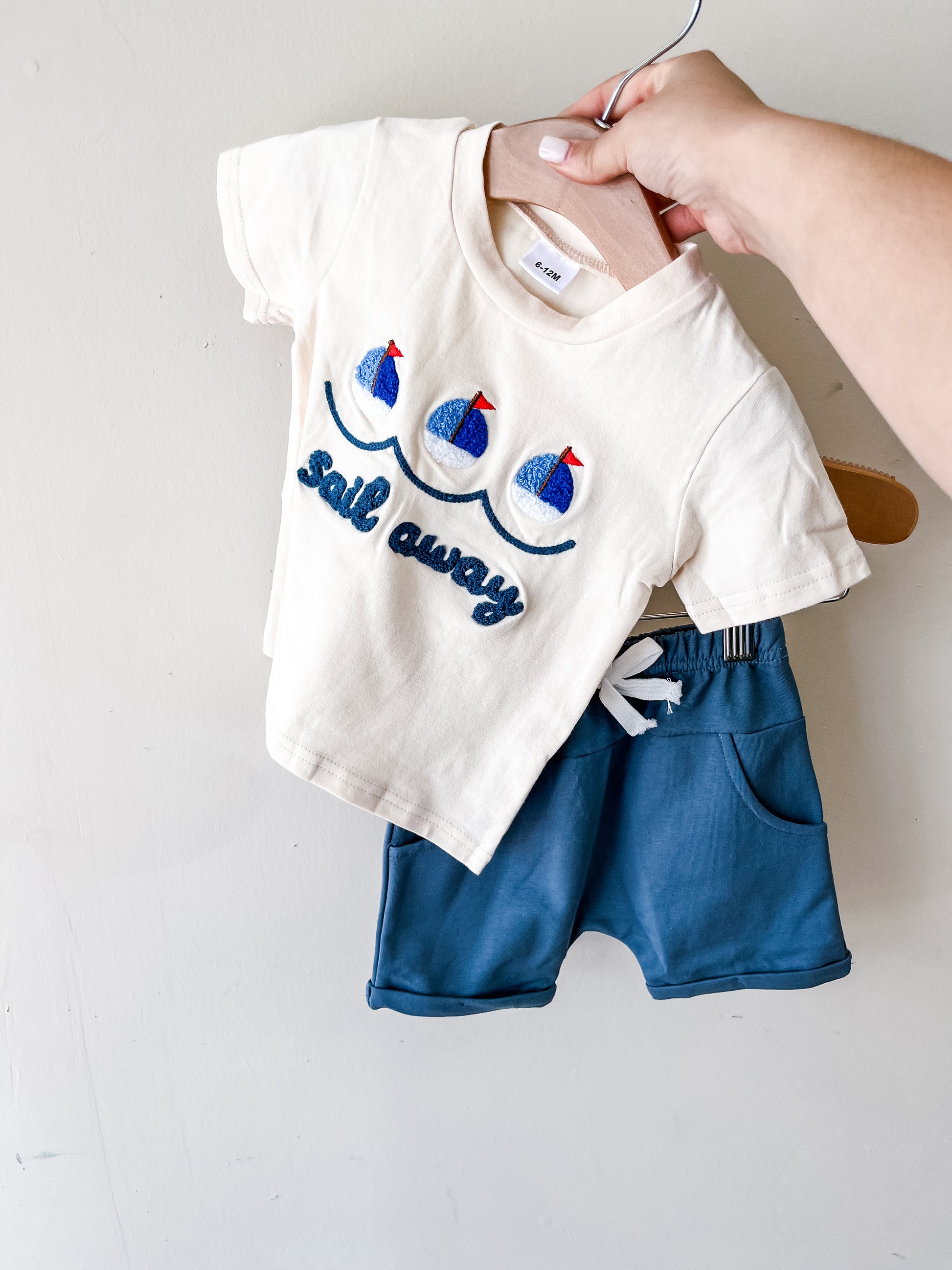 A hand holds the Tiny Tot Bundles Sail Away Tee Set: a cream t-shirt with blue embroidered sailboats and "Sail away" text, paired with blue shorts featuring a white drawstring.