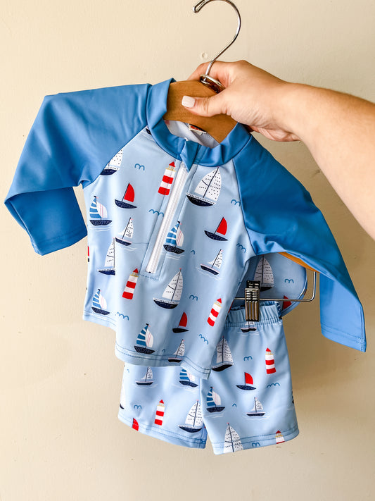 A hand holds up the Harbor Breeze Swim Set by Tiny Tot Bundles—a two-piece light blue toddler swimsuit with long sleeves, shorts, a front zipper, and a sailboat pattern in red, white, and blue.