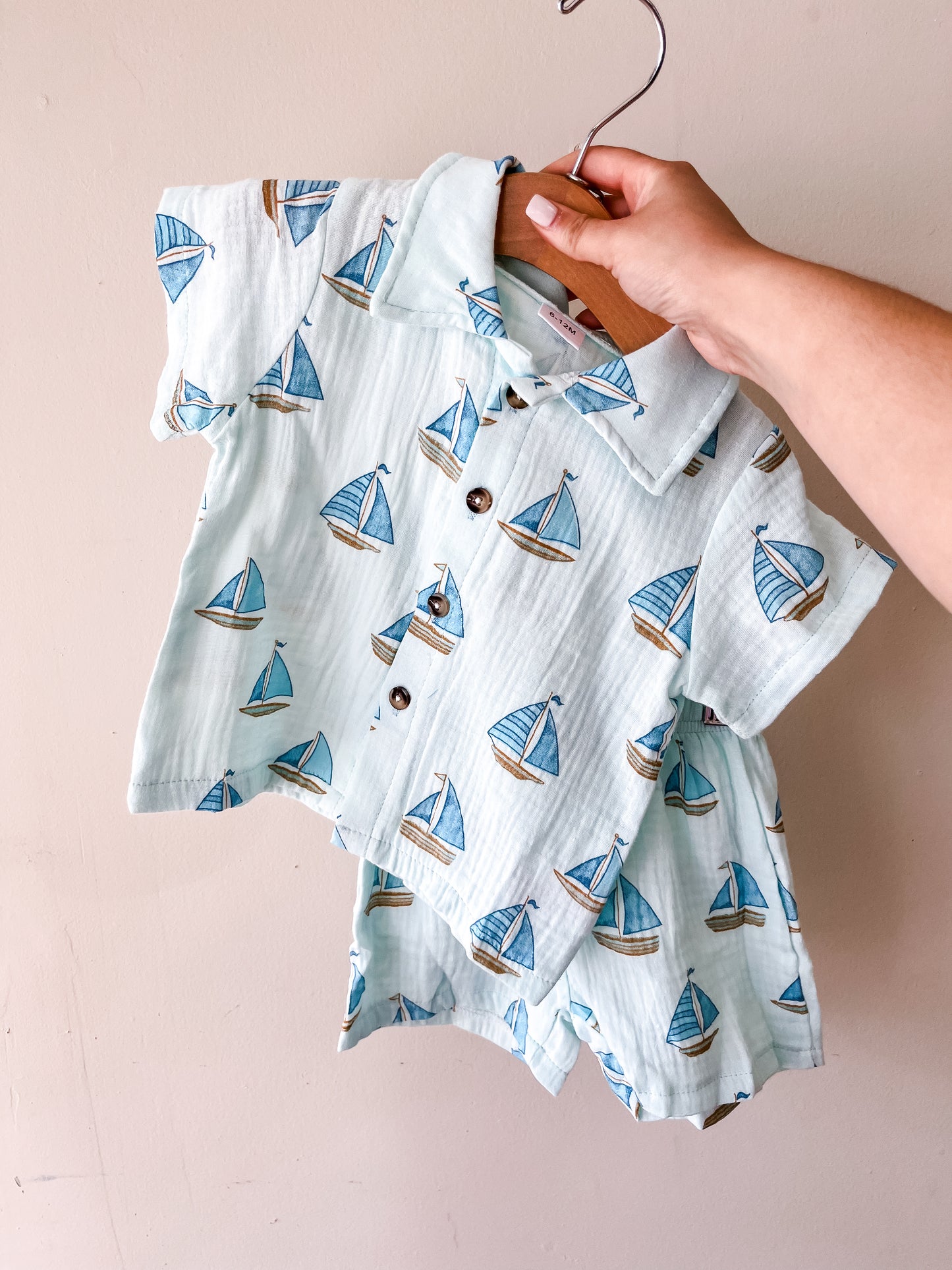 A hand displays the Tiny Tot Bundles Ocean Voyage Linen Set— a child's short-sleeve button-up shirt and matching shorts, both featuring a blue sailboat print—on a wooden hanger against a beige background.