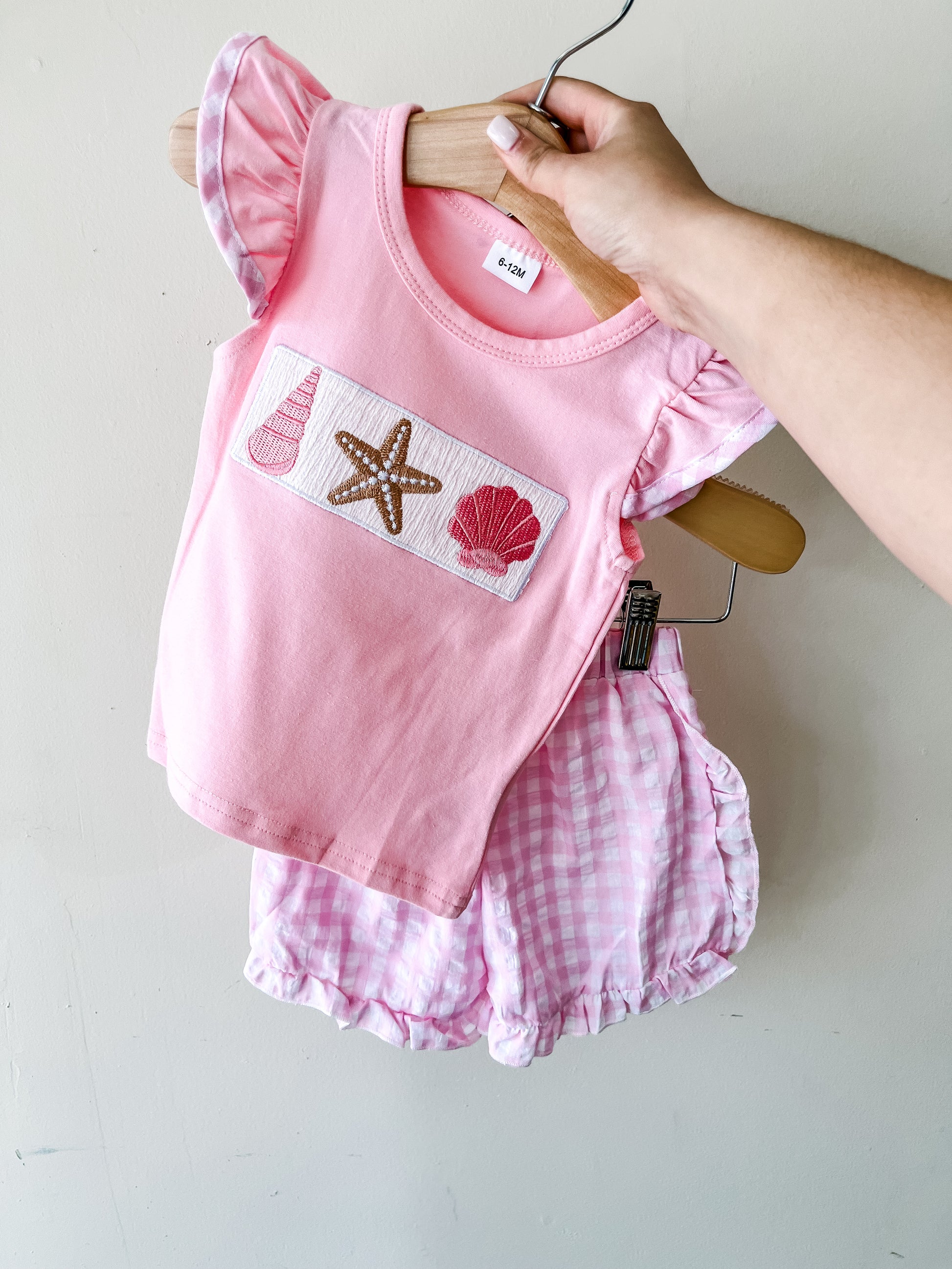 A hand holds the Tiny Tot Bundles Seaside Sweetheart Short Set, featuring a pink short-sleeve top with seashell and starfish designs and matching pink gingham shorts.