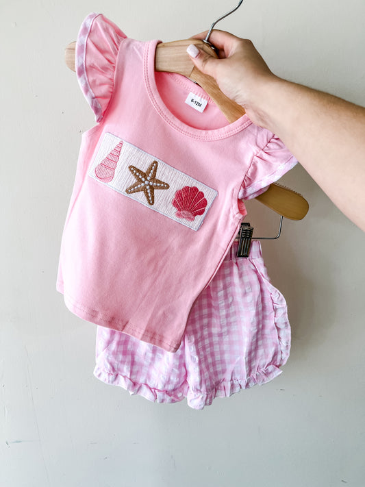 A hand holds the Tiny Tot Bundles Seaside Sweetheart Short Set, featuring a pink short-sleeve top with seashell and starfish designs and matching pink gingham shorts.