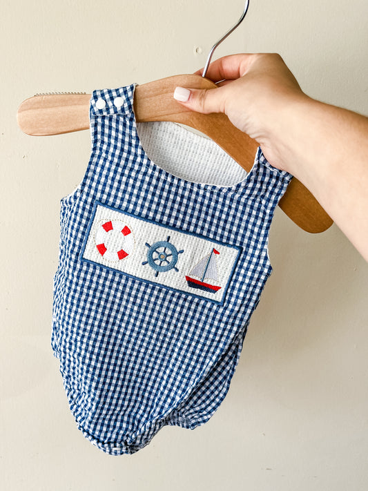 A hand holds the Tiny Tot Bundles Blue Gingham Harbor Romper on a wooden hanger, showcasing its nautical embroidery of a life preserver, ship wheel, and sailboat.