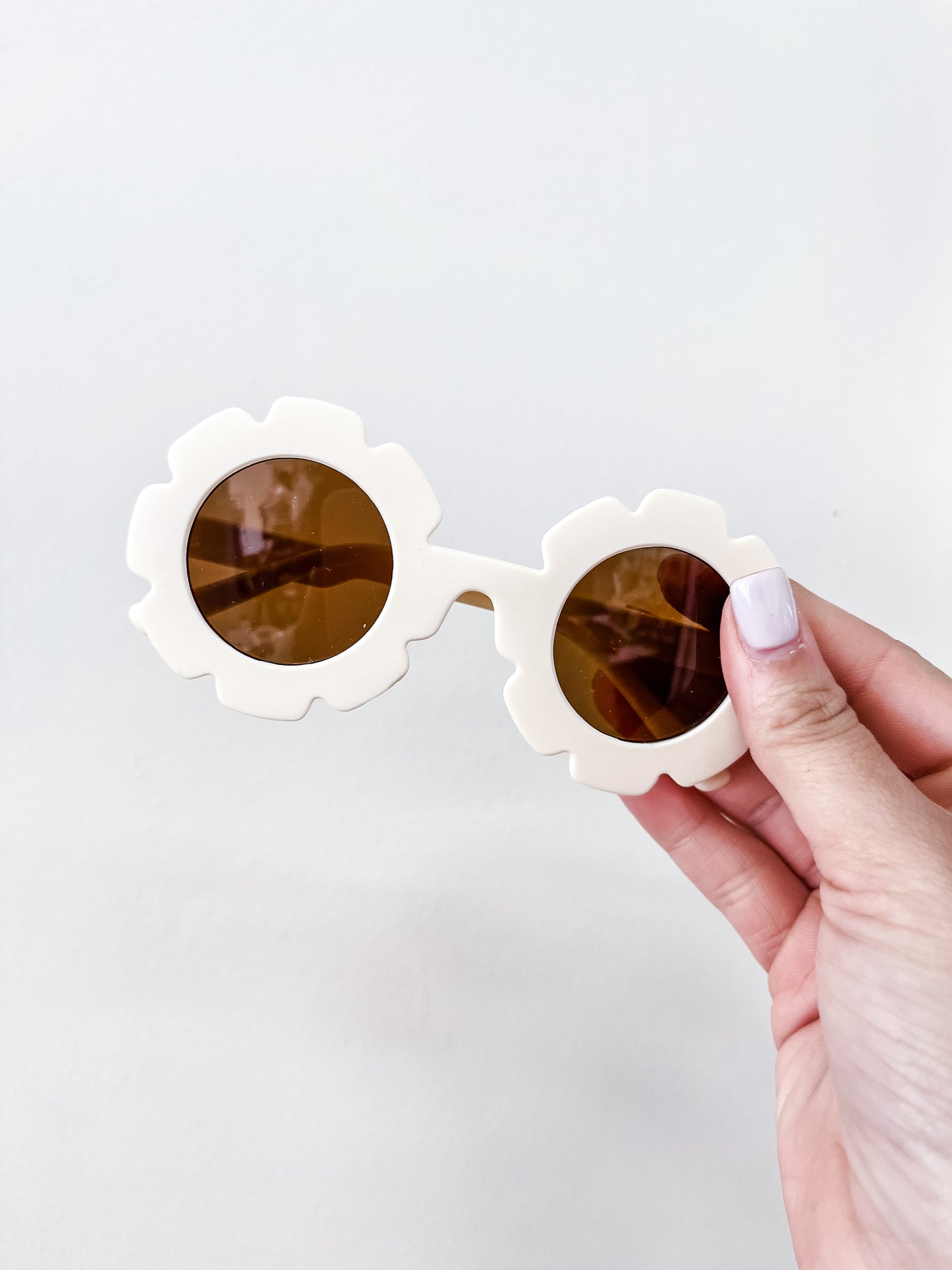 A hand with light-painted nails holds the Neutral Floral Sunglasses by Tiny Tot Bundles—round, flower-shaped frames in white with brown lenses—against a plain white background.