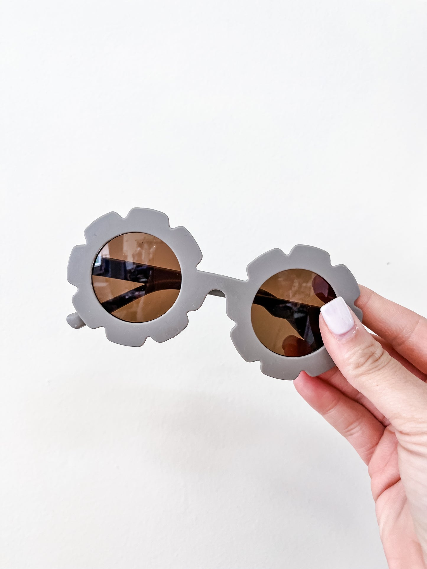 A hand with light pink nails holds Tiny Tot Bundles' Neutral Floral Sunglasses, featuring round, flower-shaped gray frames and brown tinted lenses, against a plain white background.