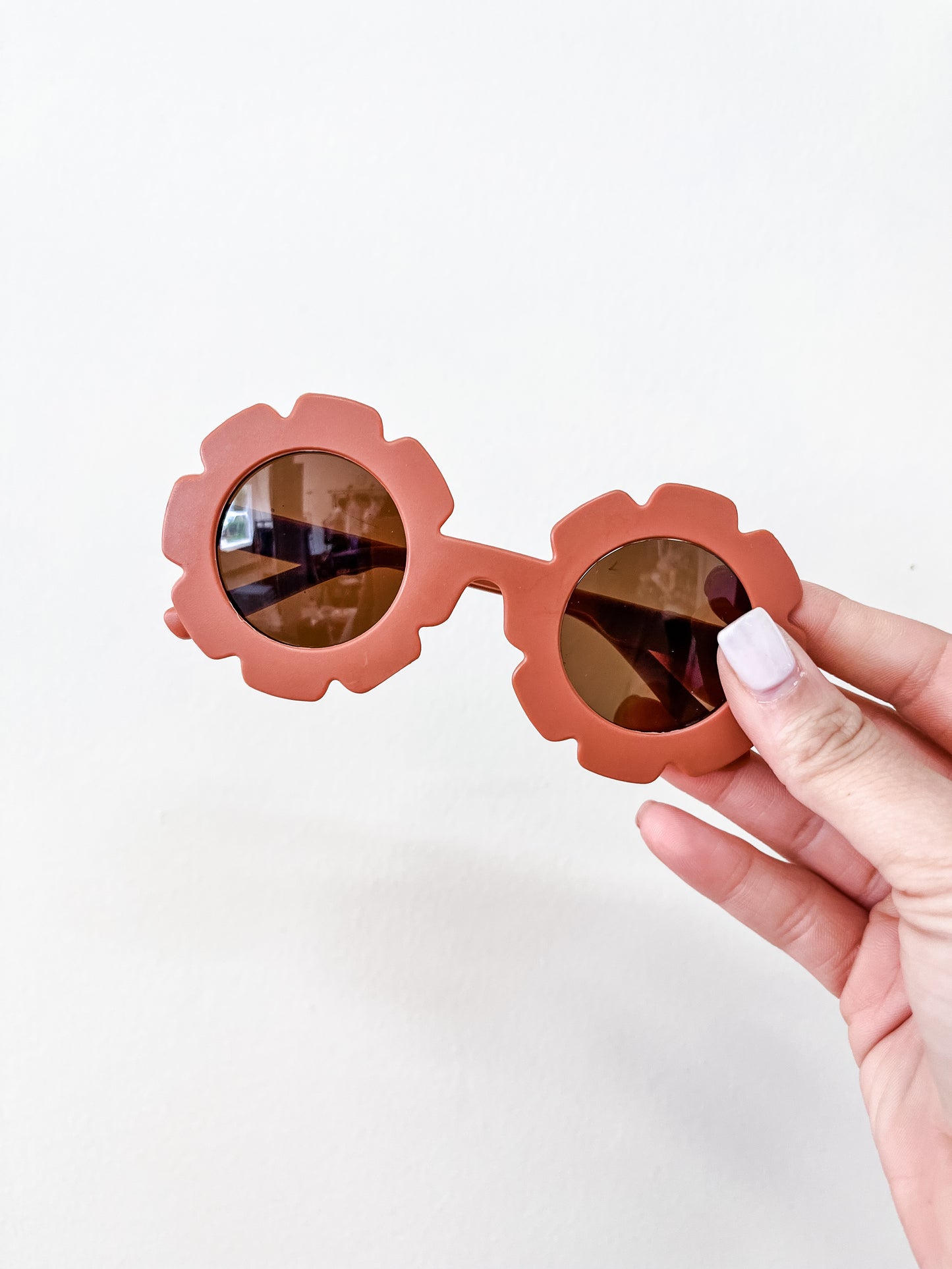 A hand with light pink nail polish holds the Neutral Floral Sunglasses by Tiny Tot Bundles, featuring a brown flower-shaped frame, against a plain white background.