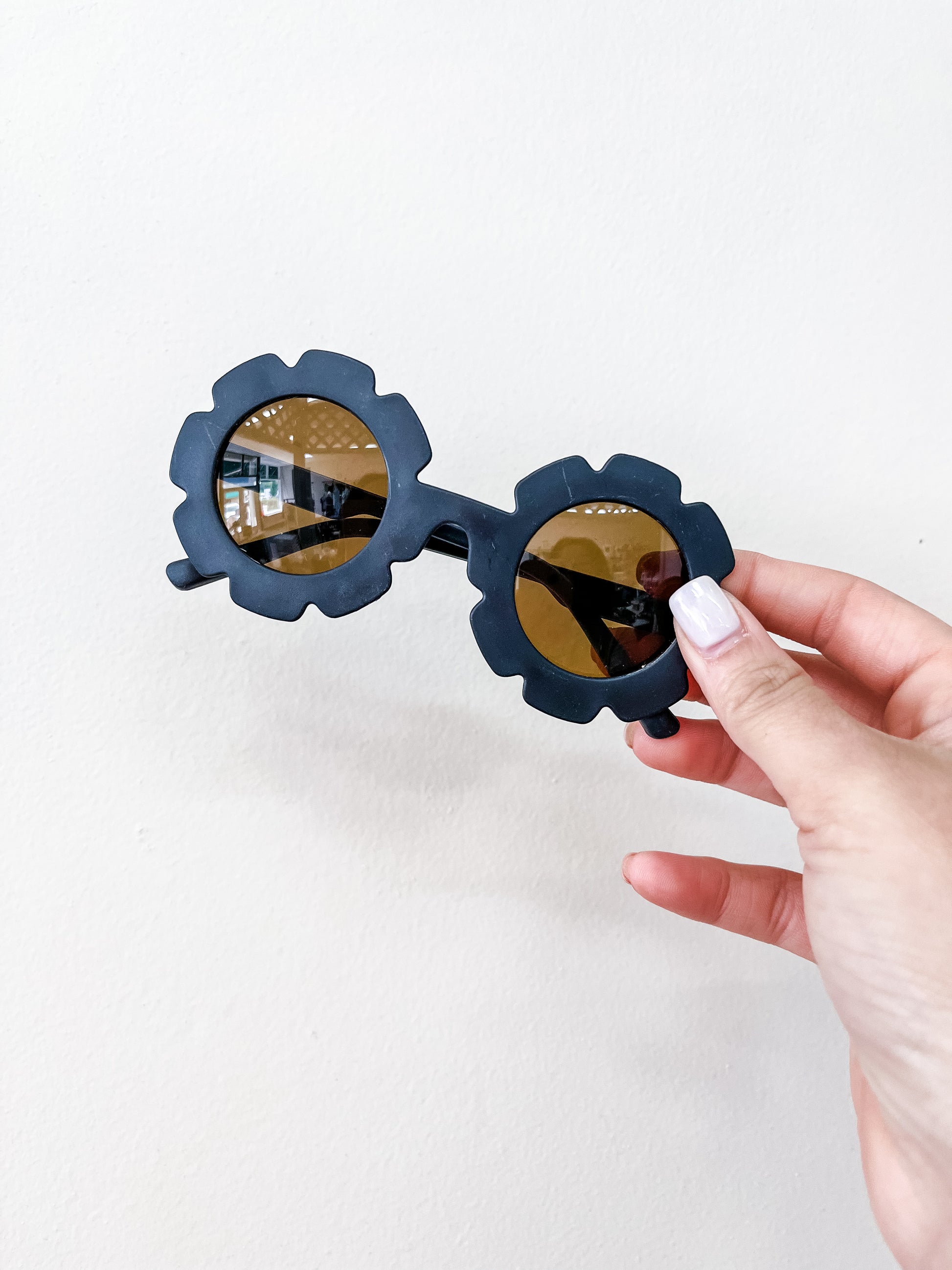 A hand with light pink nail polish holds Tiny Tot Bundles' Neutral Floral Sunglasses, featuring black flower-shaped frames and round amber lenses, against a plain white background.