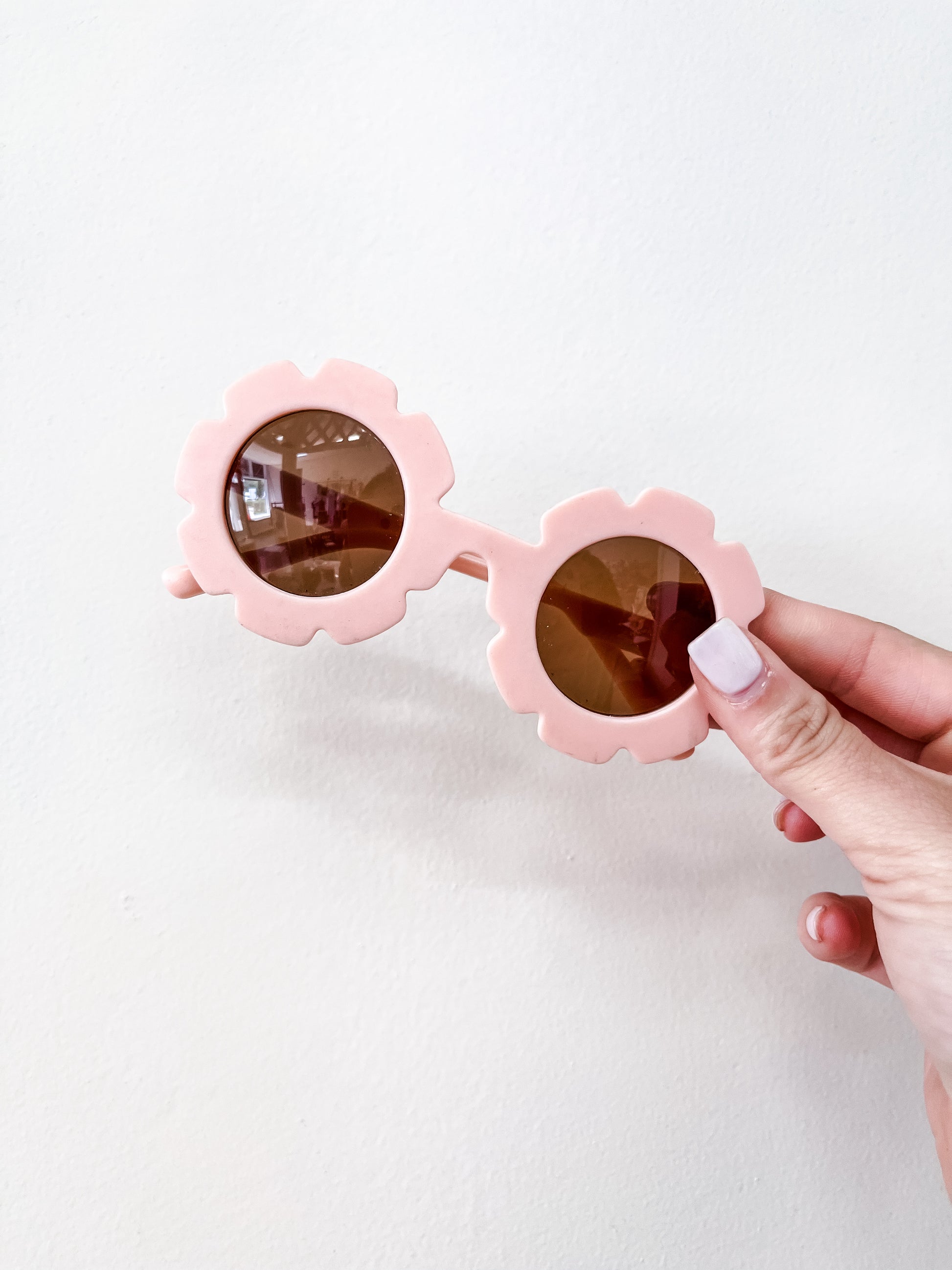 A hand with light pink nail polish holds the Tiny Tot Bundles Neutral Floral Sunglasses, featuring round, light pink, flower-shaped frames, against a plain white background.
