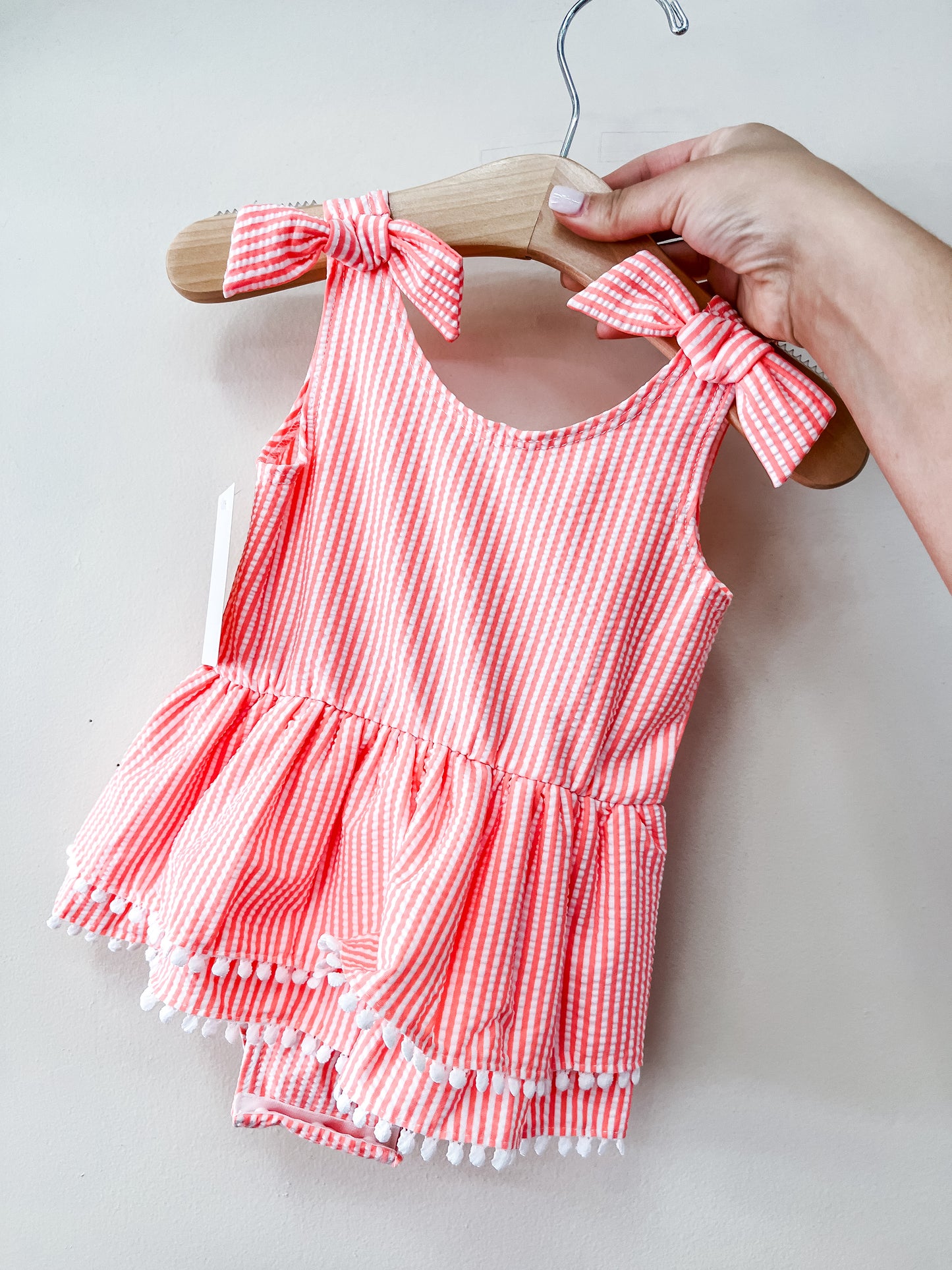 A hand holds a wooden hanger with the Tiny Tot Bundles Coral Coast Swimsuit—a pink and white striped baby swimsuit with shoulder bows, a ruffled peplum hem, white pom-pom trim, and matching bloomers.
