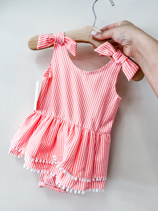 A hand holds a wooden hanger with the Tiny Tot Bundles Coral Coast Swimsuit—a pink and white striped baby swimsuit with shoulder bows, a ruffled peplum hem, white pom-pom trim, and matching bloomers.