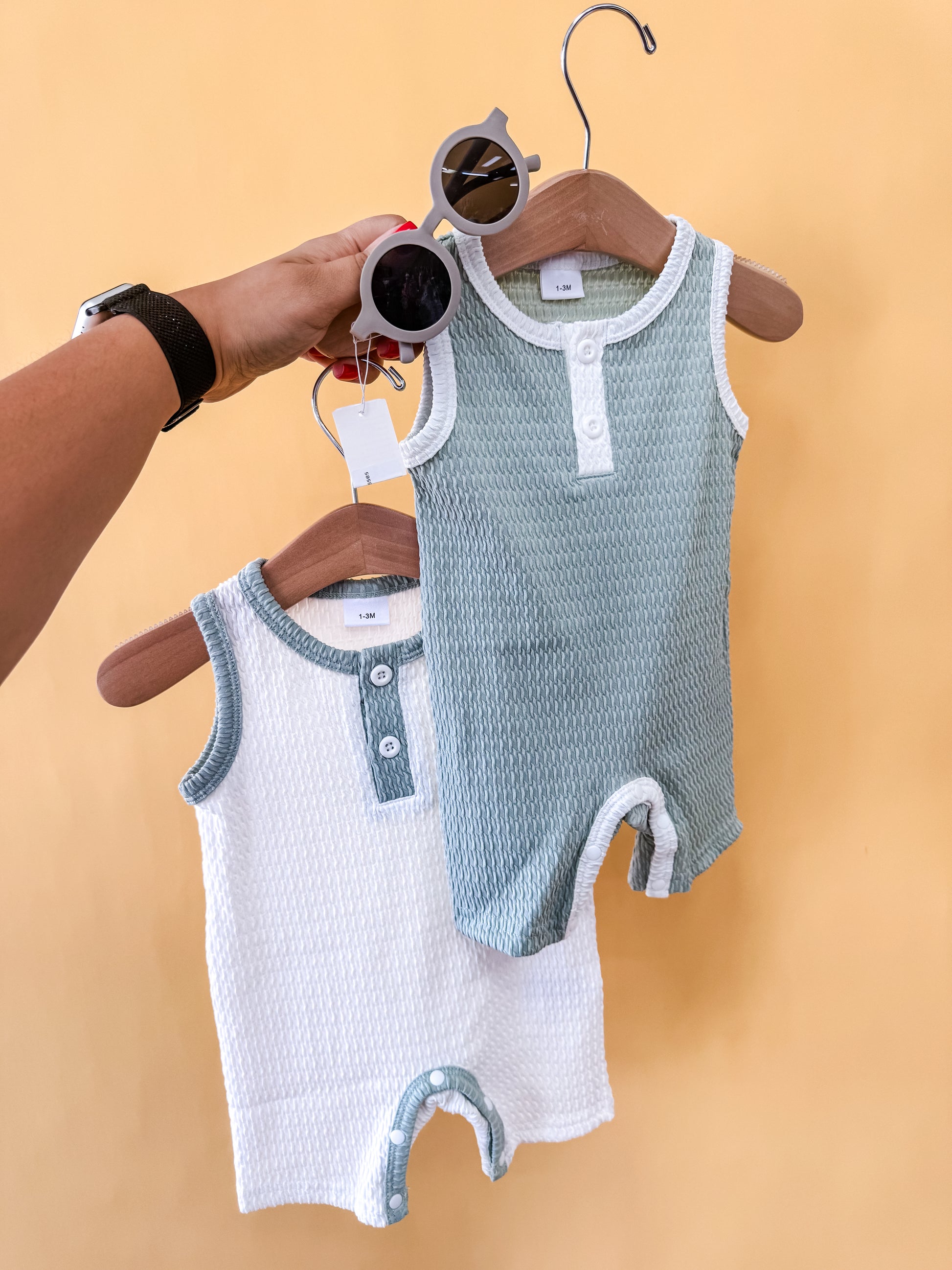 A hand holds a hanger with baby sunglasses and a green sleeveless romper from Tiny Tot Bundles' "Bundle Of The Day - Boys" set, next to a white and green sleeveless romper, all against a yellow background.