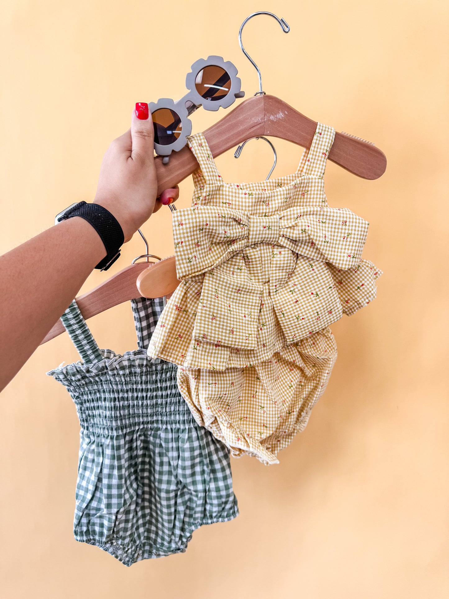 A hand displays Tiny Tot Bundles' "Bundle Of The Day - Girl": a yellow romper with a bow, a green and white checkered romper, and flower-shaped sunglasses—all set against a solid light yellow background.
