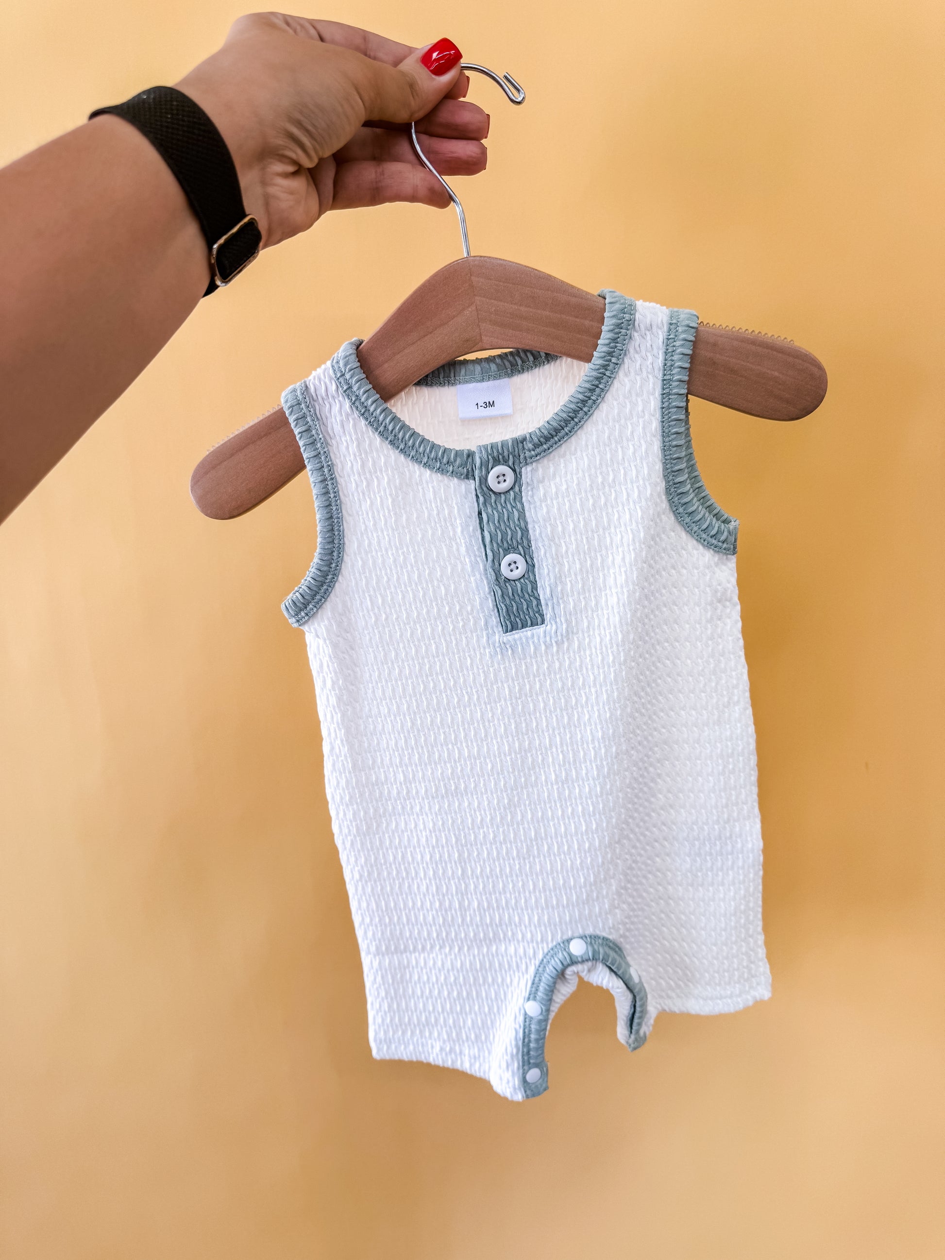 A hand holds a wooden hanger displaying the Tiny Tot Bundles Harbor Mist Romper, a sleeveless white baby outfit with light blue trim and two buttons, set against a plain yellow background.