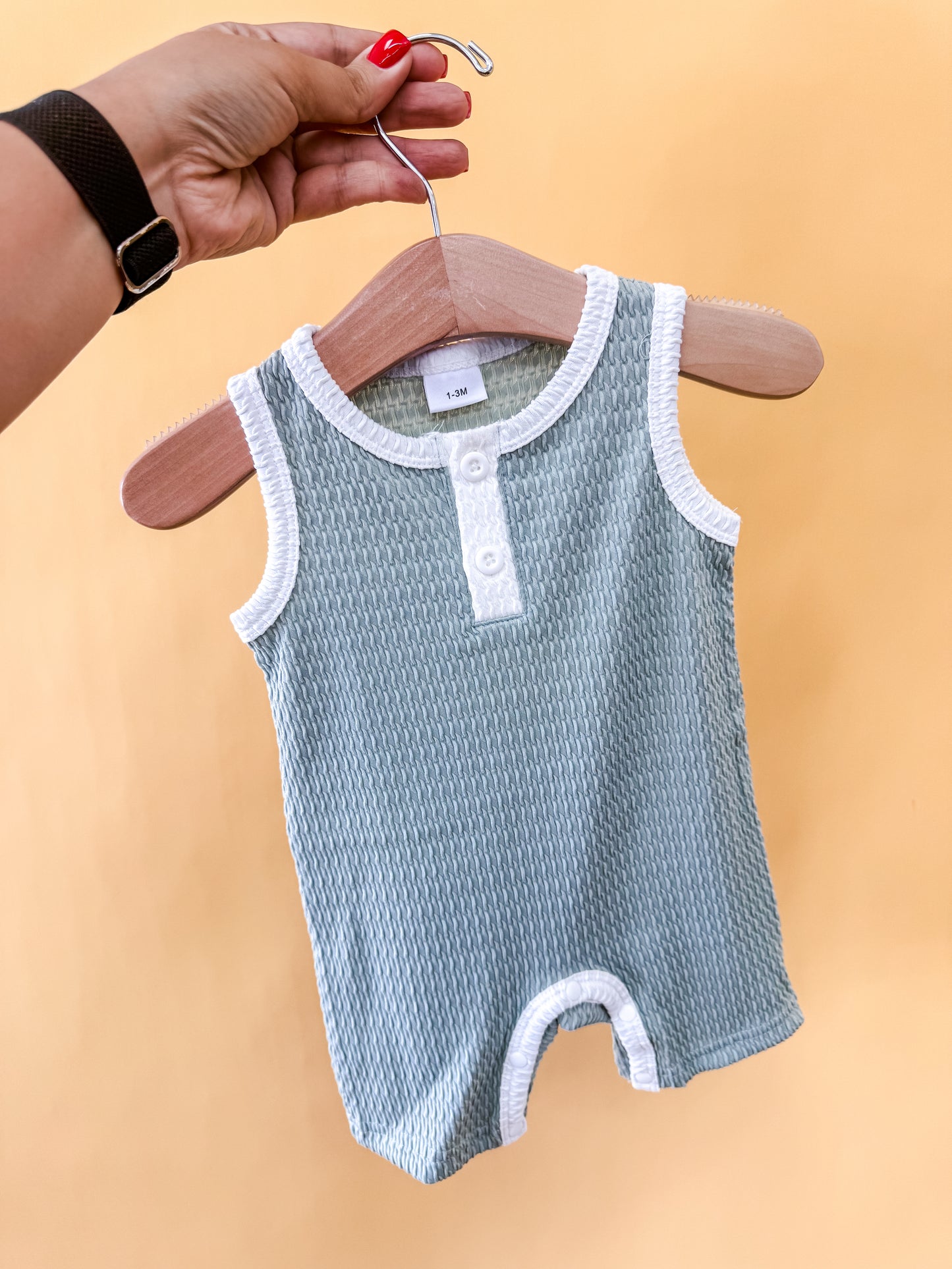 A hand with red nail polish holds a hanger displaying the Tiny Tot Bundles Sage Harbor Romper, a sleeveless, light blue textured baby outfit with white trim and buttons, set against a plain yellow background.