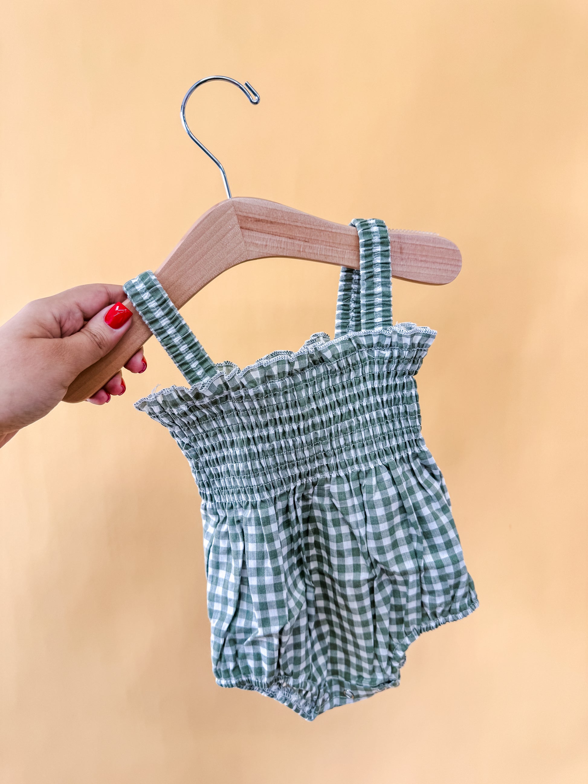 A hand with red nail polish holds a wooden hanger displaying the Meadow Mist Romper by Tiny Tot Bundles, a green and white checkered baby romper with ruffled trim and suspenders, set against a plain light yellow background.