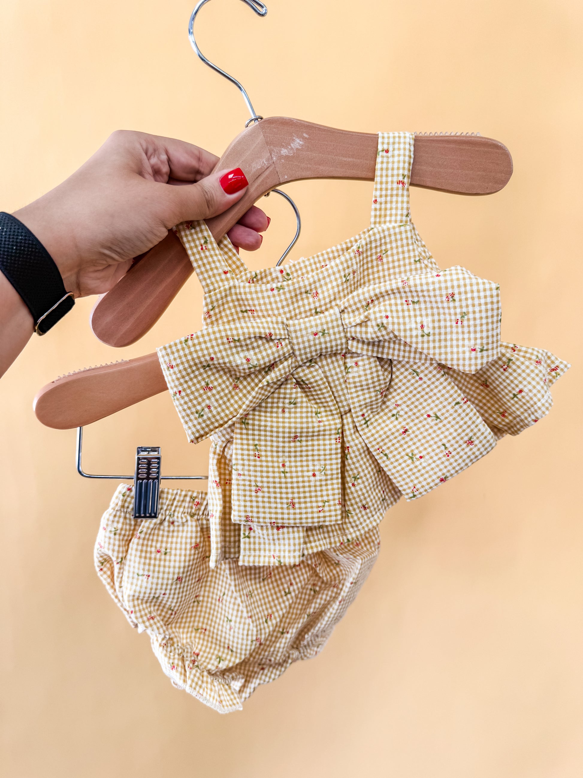 A hand holds Tiny Tot Bundles' Buttercup Bloom Set—a baby girl's yellow plaid outfit with a big bow top and matching bloomers—against a peach background.