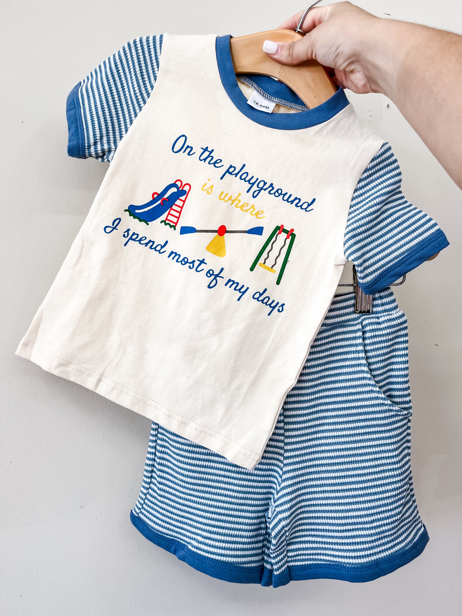 A hand holds the Tiny Tot Bundles Playground Day's Set, a blue and white striped toddler outfit with a shirt that reads "On the playground is where I spend most of my days" and features playground equipment illustrations.