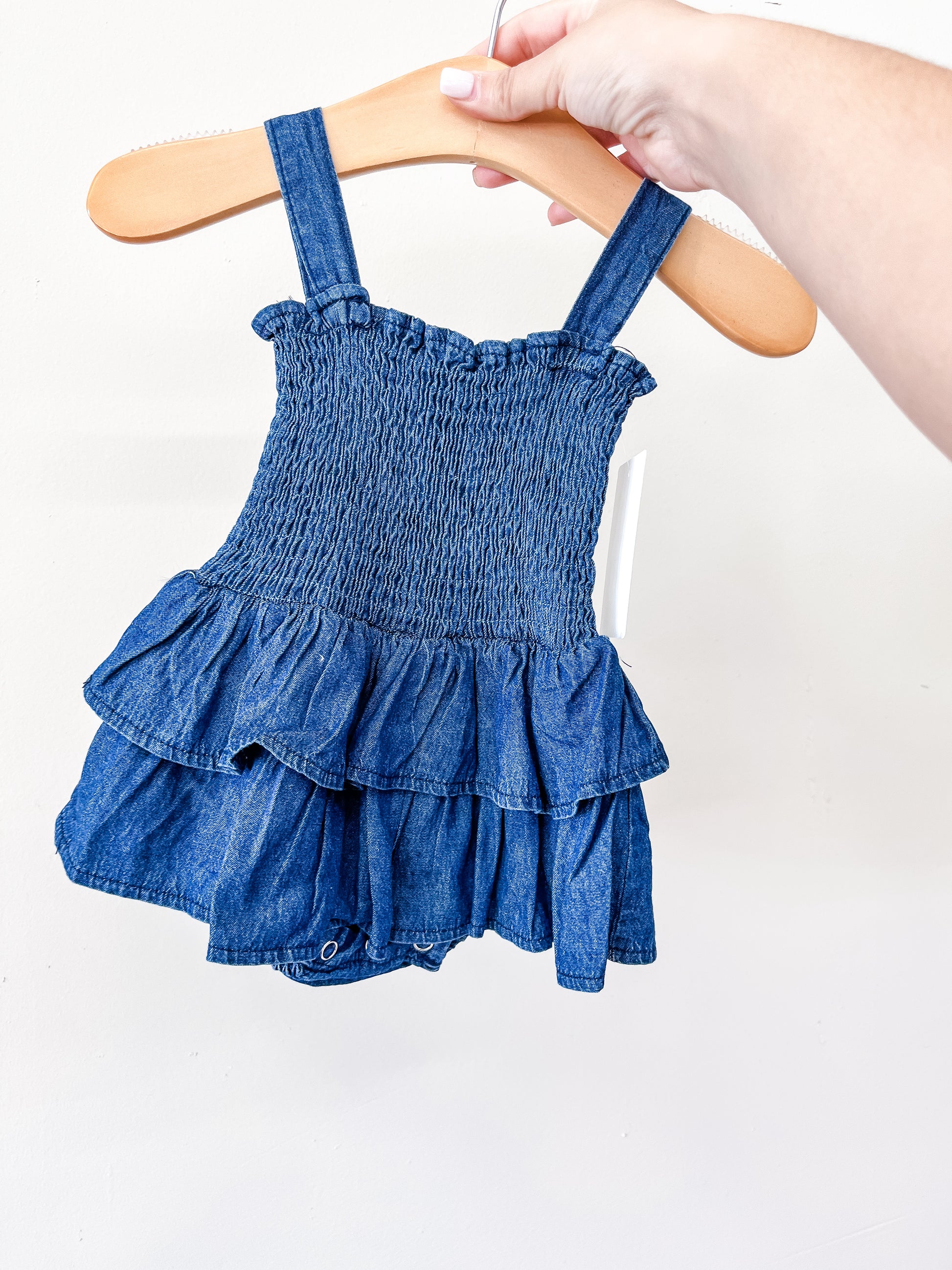 A hand holds a wooden hanger displaying the Tiny Tot Bundles Denim Ruffle Romper, featuring a baby blue shade, shoulder straps, and a smocked bodice, shown against a plain white background.