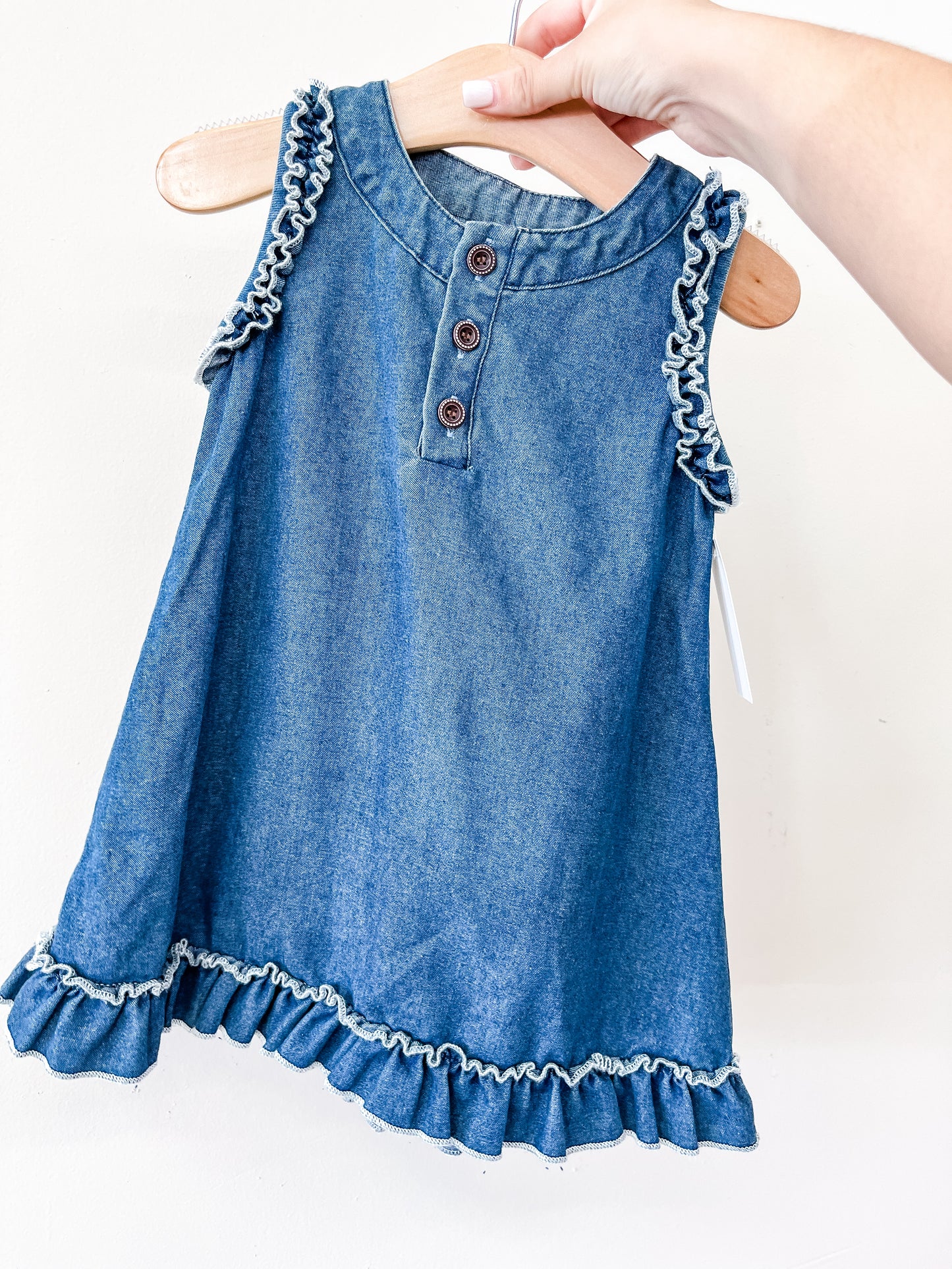 A hand holds the Tiny Tot Bundles Denim Ruffle Dress, a blue denim sleeveless baby dress with ruffled shoulders and hem, and three brown front buttons, displayed on a wooden hanger.