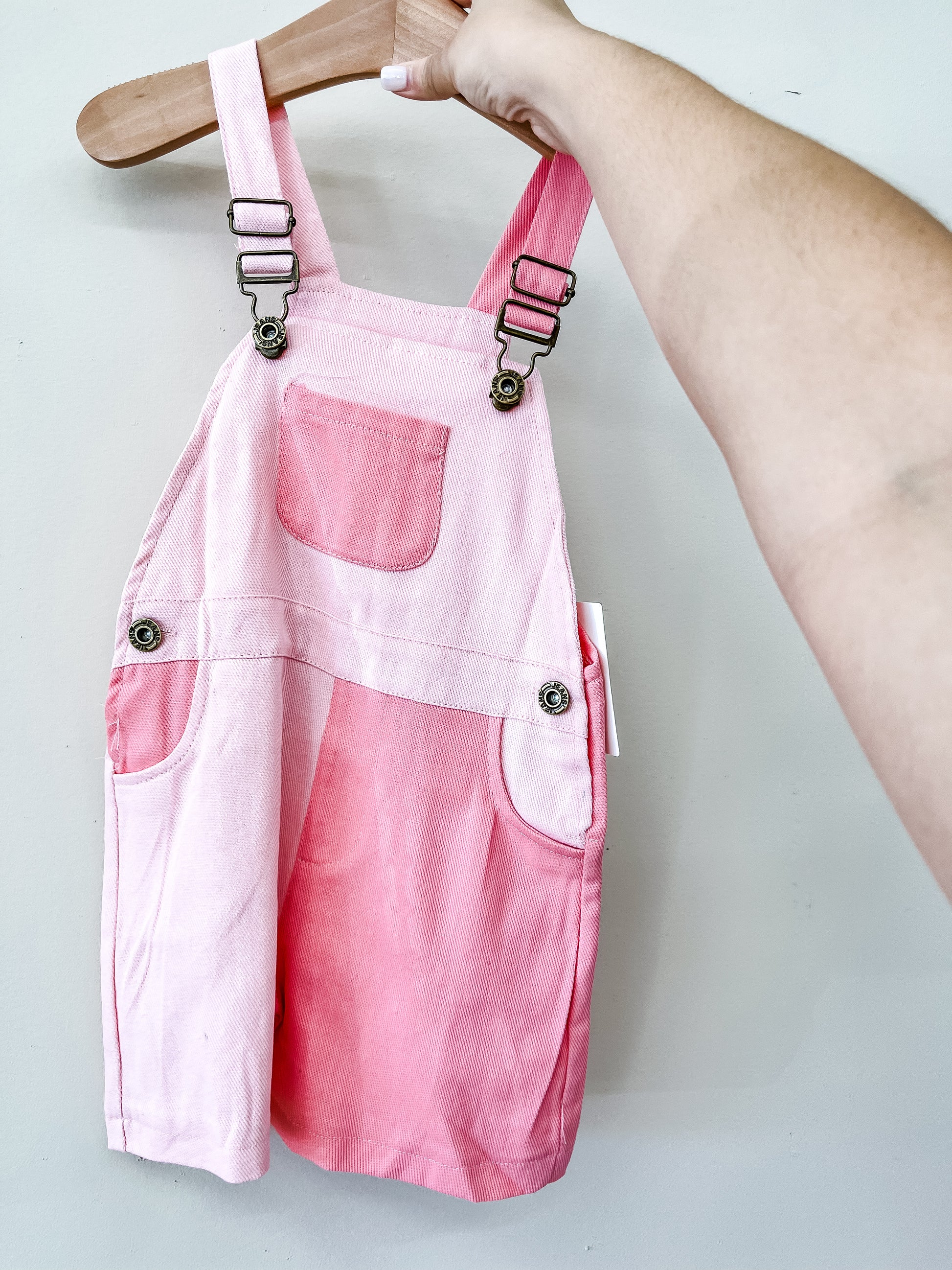 A hand holds up the Tiny Tot Bundles Pink Color Block Overalls on a wooden hanger against a light gray background, featuring metal clasps, a center chest pocket, and two front pockets.