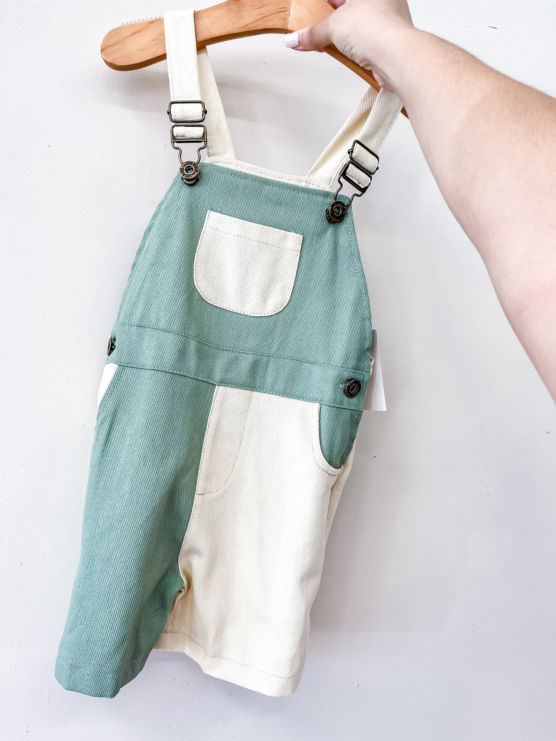 The Tiny Tot Bundles Green Color Block Overalls, featuring a front pocket, are displayed on a wooden hanger against a plain white background.