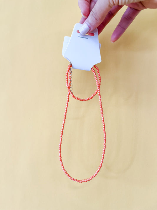 A hand with white-painted nails holds a Tiny Tot Bundles Summer Beaded Bracelet Set featuring small, bright orange beads and a gold chain clasp, displayed on a white card against a light yellow background.