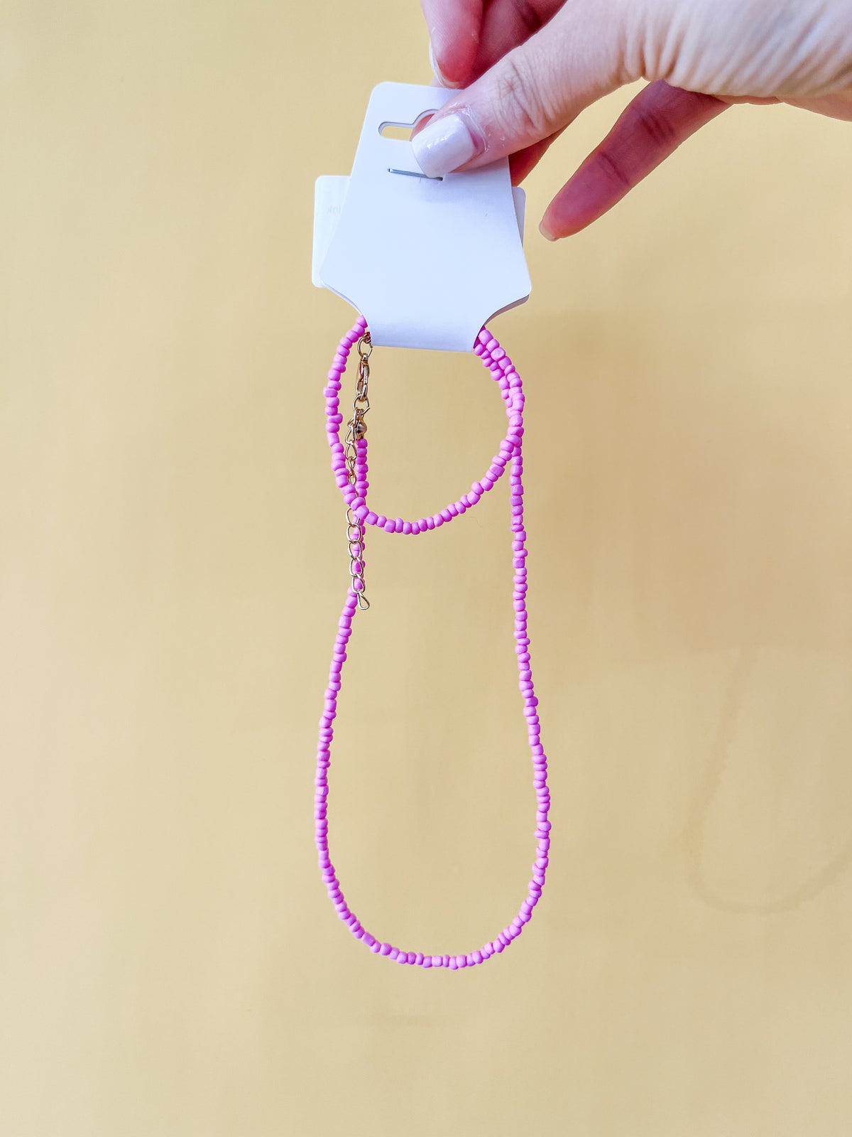 A hand with a light pink manicure holds a white card showing the Tiny Tot Bundles Summer Beaded Bracelet Set—small bright pink beads with a gold clasp—against a solid yellow background.