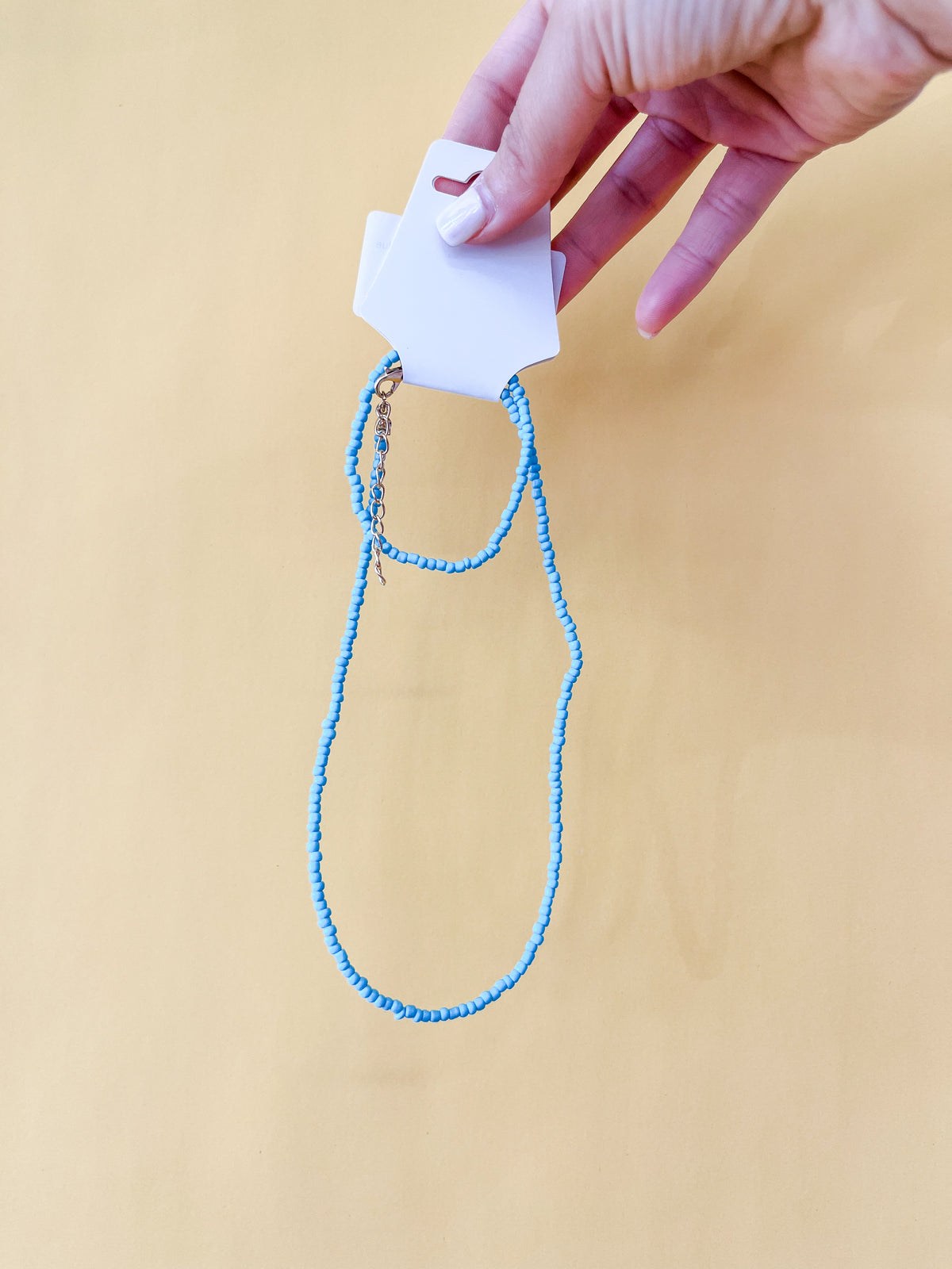 A hand with white nail polish holds a white jewelry card displaying the Tiny Tot Bundles Summer Beaded Bracelet Set with small blue beads against a pale yellow background.