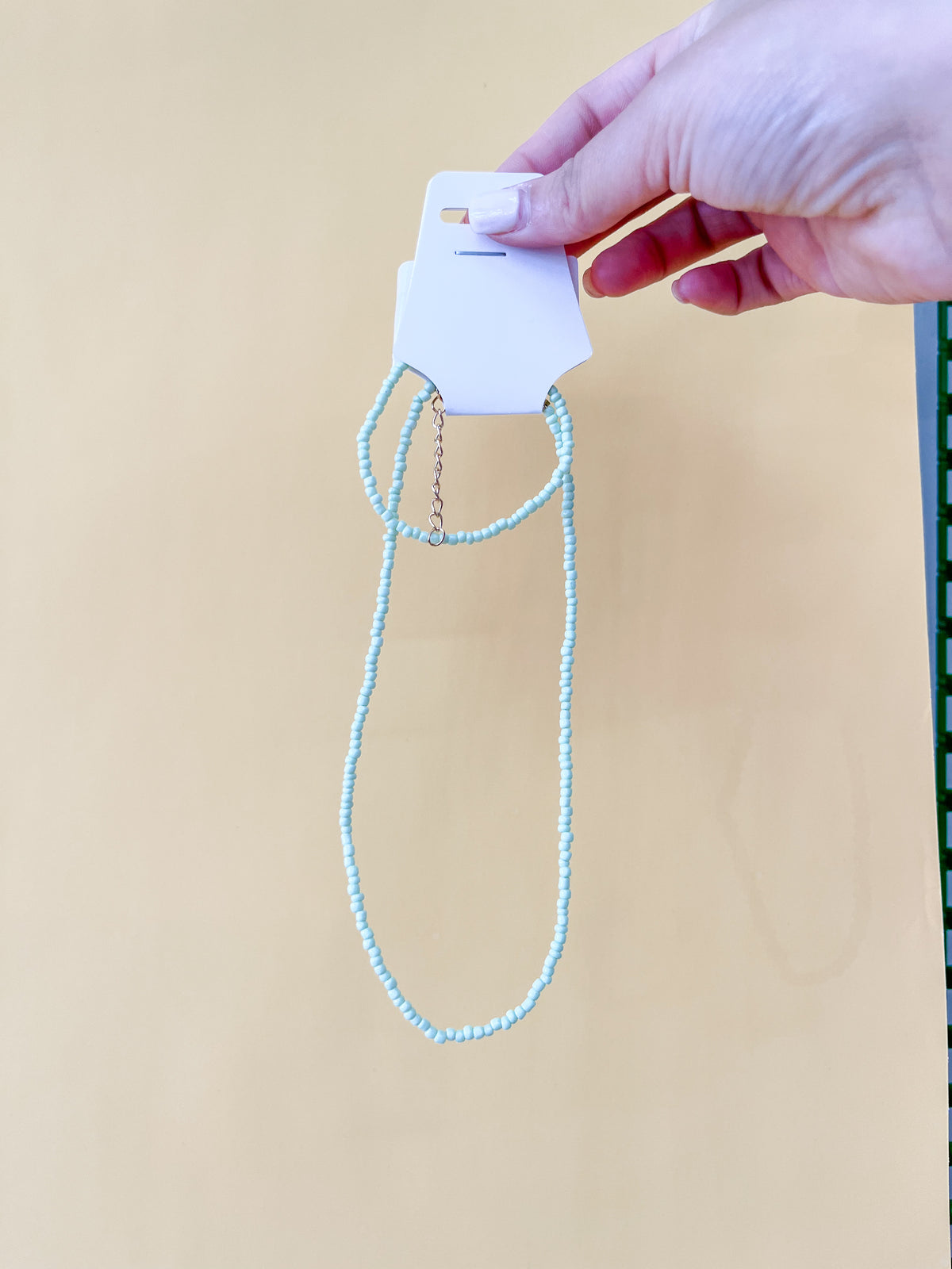 A hand with light-colored nails holds a card displaying the Tiny Tot Bundles Summer Beaded Bracelet Set, featuring delicate light blue beads and a thin gold chain extension, set against a beige background.