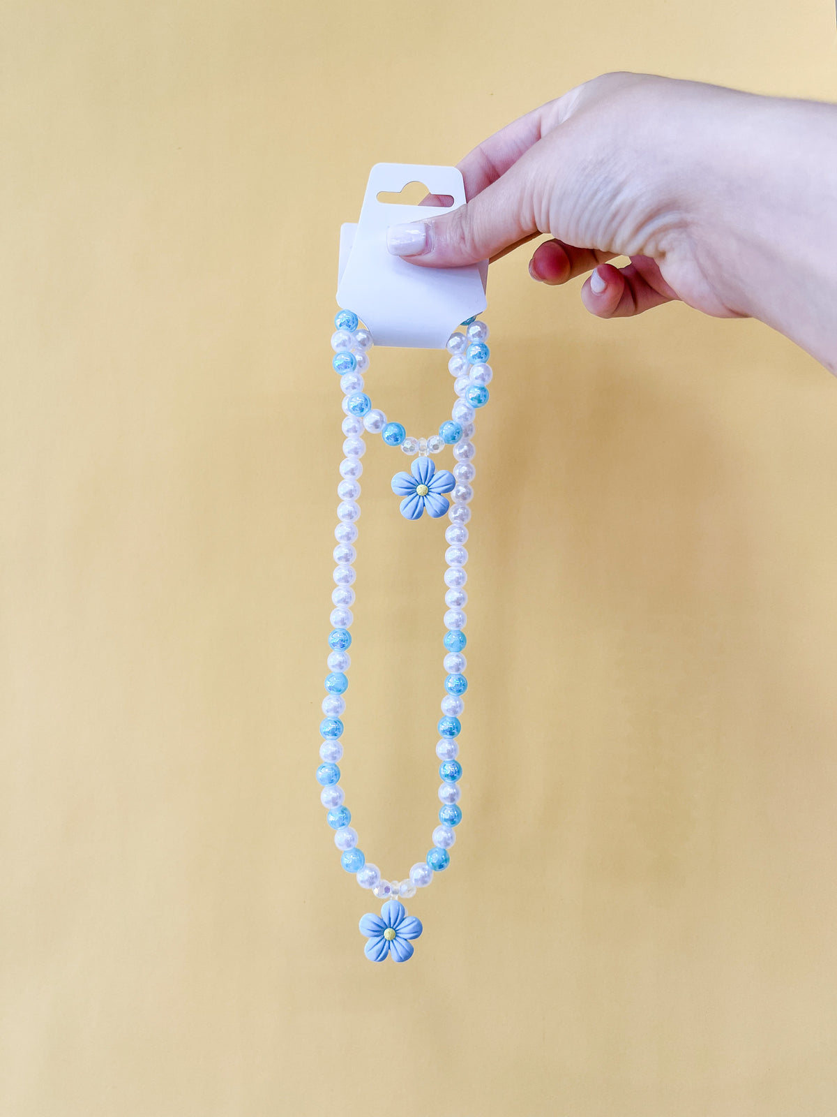 A hand holds a Tiny Tot Bundles Pearl Daisy Necklace Set, featuring two beaded necklaces with blue and white beads and matching flower charms, against a plain yellow background.
