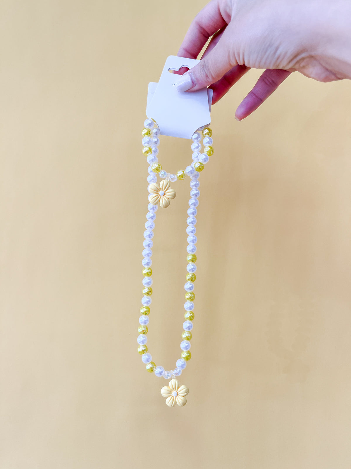A hand holds a card displaying the Pearl Daisy Necklace Set by Tiny Tot Bundles, featuring a beaded necklace and bracelet with yellow and white beads and yellow flower charms, against a light yellow background.