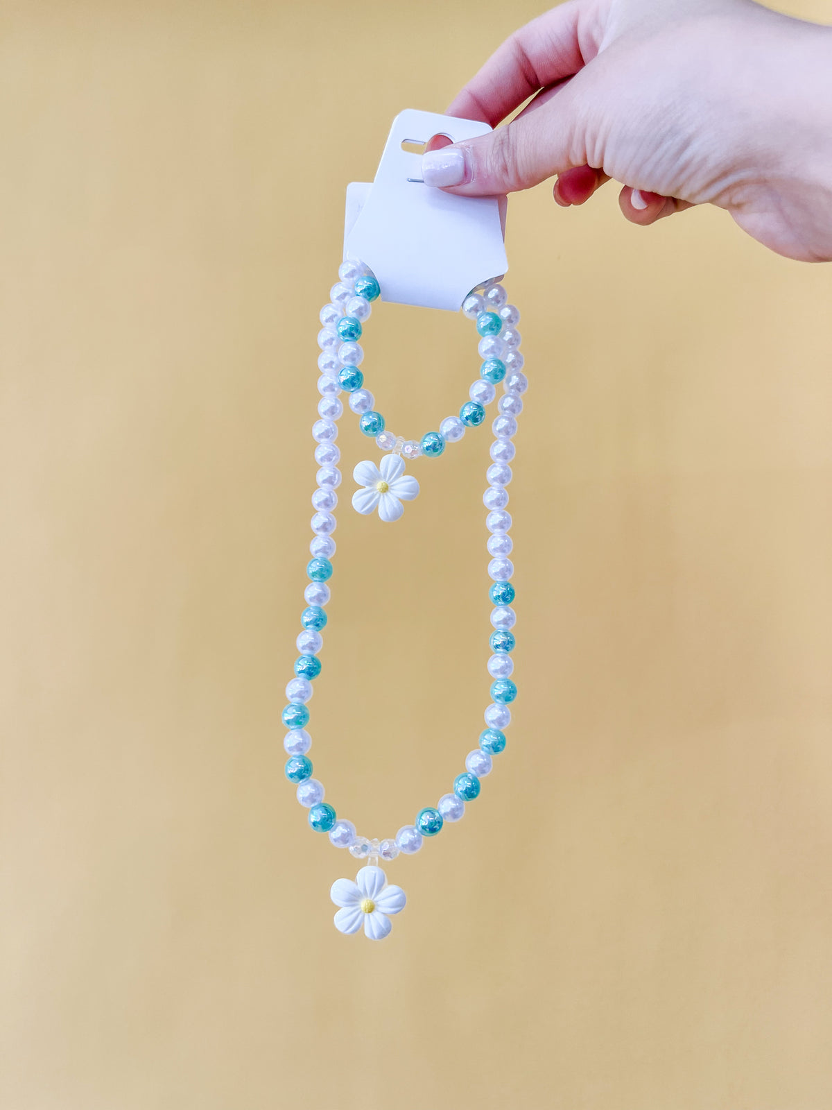 A hand displays the Tiny Tot Bundles Pearl Daisy Necklace Set, featuring two beaded necklaces with blue and white beads and daisy flower pendants, against a plain yellow background.