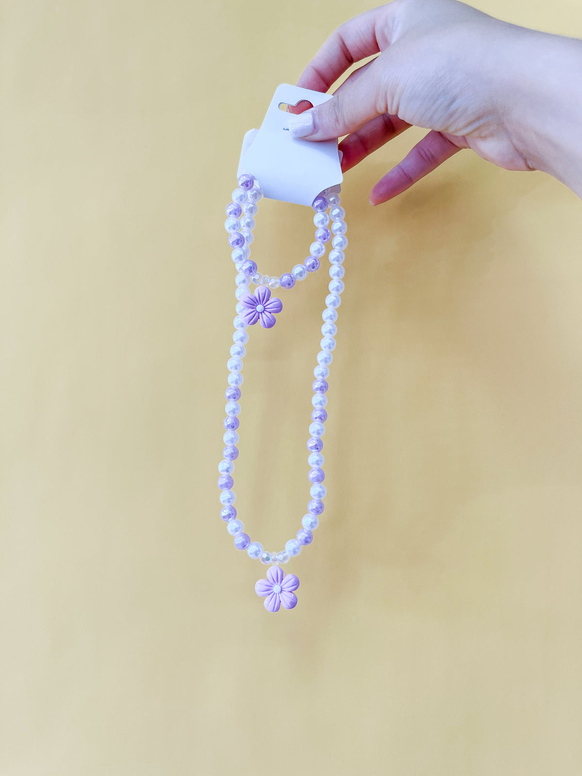 A hand with painted nails holds a Tiny Tot Bundles Pearl Daisy Necklace Set—purple and white beads with a purple flower pendant—displayed on a card against a yellow background.