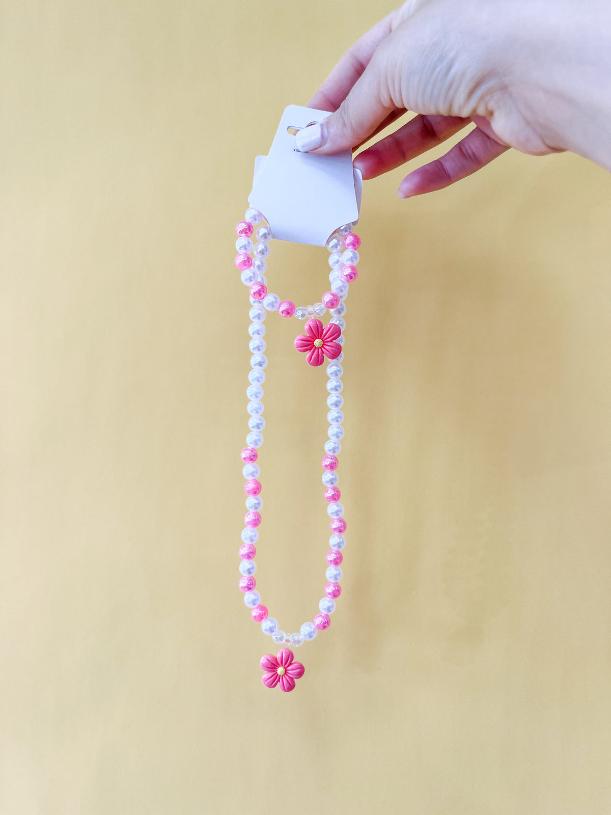 A hand displays the Tiny Tot Bundles Pearl Daisy Necklace Set, featuring pink, white, and purple beads with pink flower charms, against a plain yellow background.