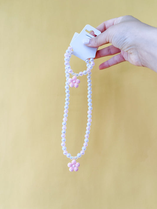 A hand holds the Tiny Tot Bundles Pearl Daisy Necklace Set—matching beaded necklace and bracelet with pink flower pendants—against a plain yellow background.