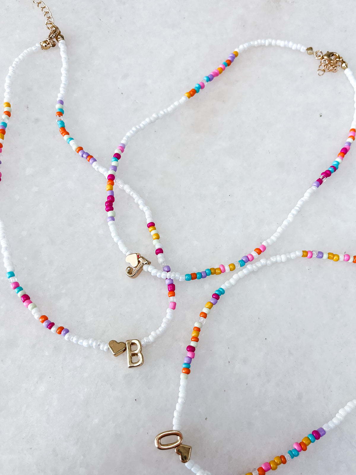 The Colorful Beaded Initial Necklace by Tiny Tot Bundles features three necklaces with colorful and white beads, each adorned with a gold heart charm and either the letter B, O, or the number 1, all displayed on a white background.